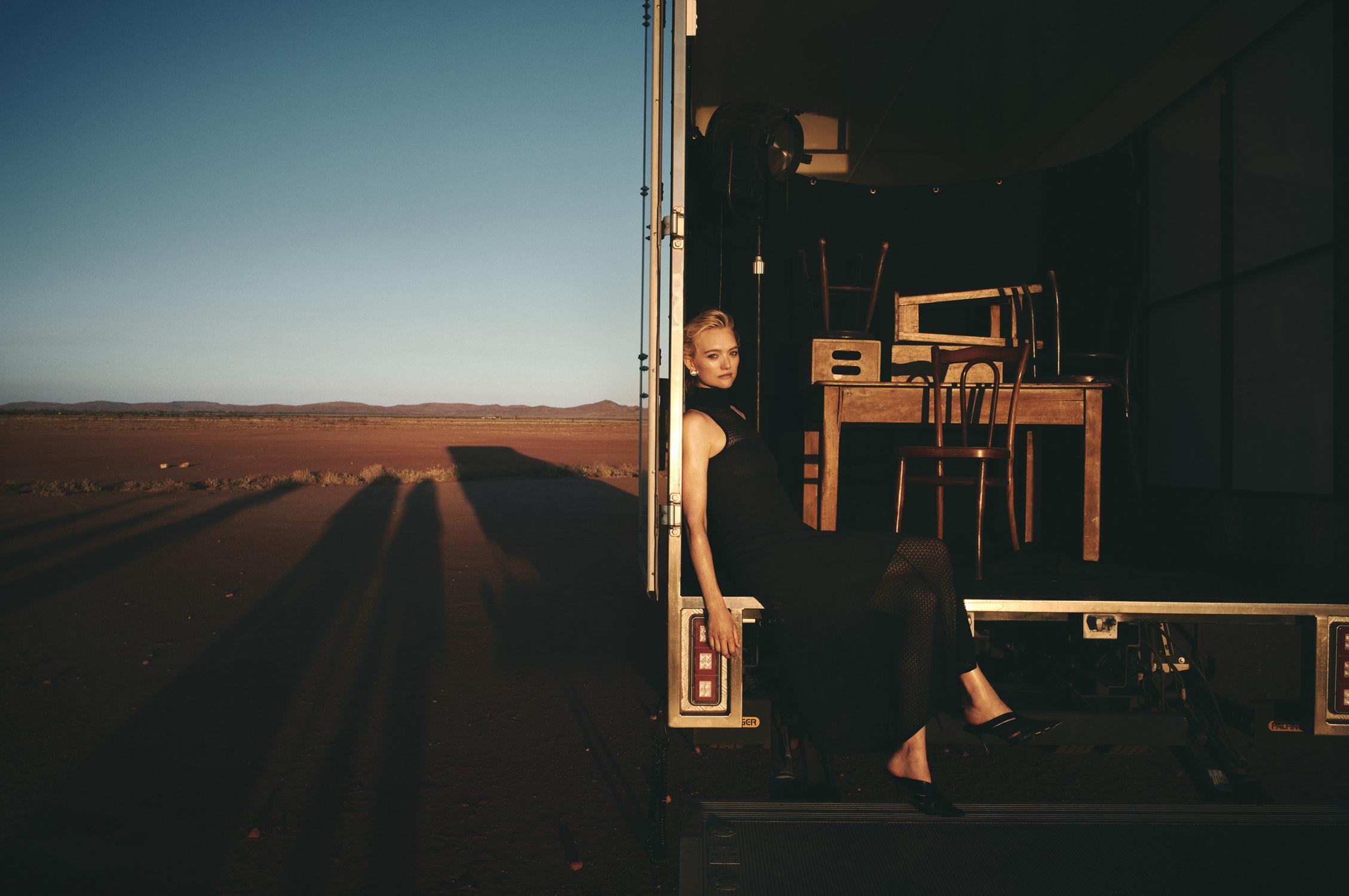 A blonde woman in a black dress poses on the edge of a trailer at dusk in the desert, with the horizon in the background.