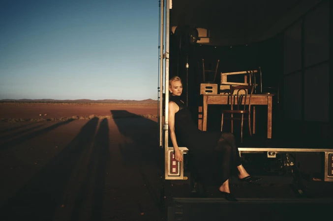 A blonde woman in a black dress poses on the edge of a trailer at dusk in the desert, with the horizon in the background.