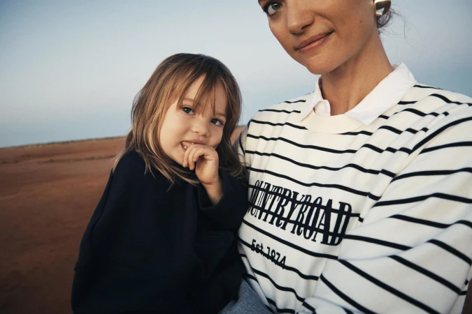 A child and an adult woman pose together outdoors in matching Country Road sweatshirts, set against a neutral desert landscape under a clear sky.