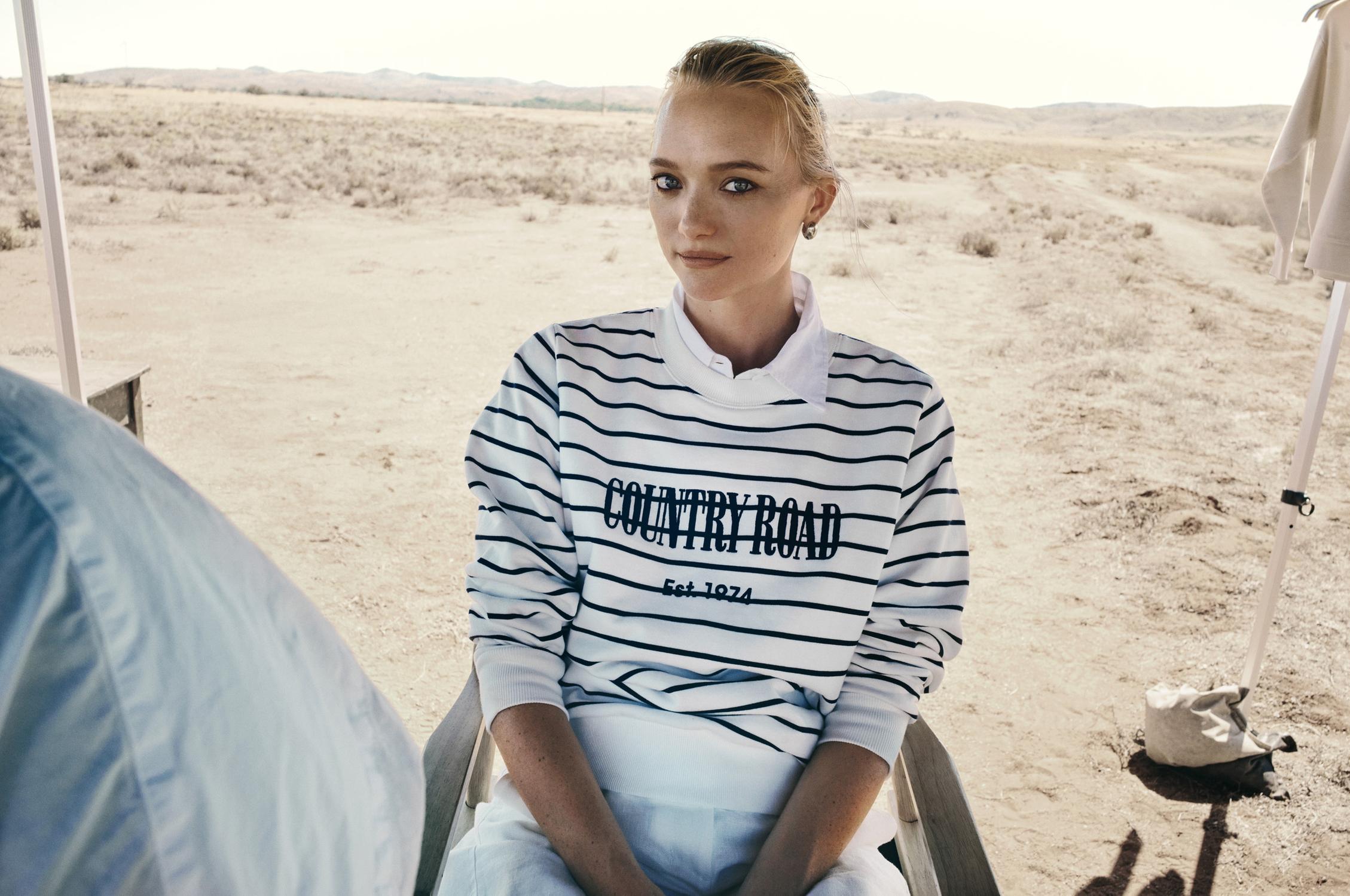 A blonde woman poses in a striped sweater with "COUNTRY ROAD" text, sitting in a chair in a sparse, sunny, desert landscape.