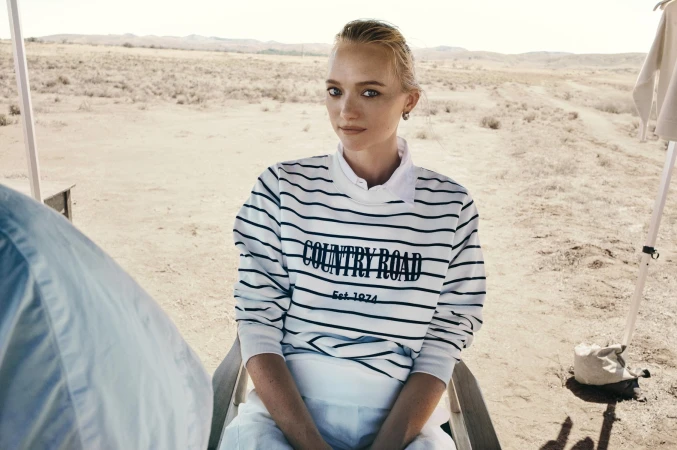 A blonde woman poses in a striped sweater with "COUNTRY ROAD" text, sitting in a chair in a sparse, sunny, desert landscape.