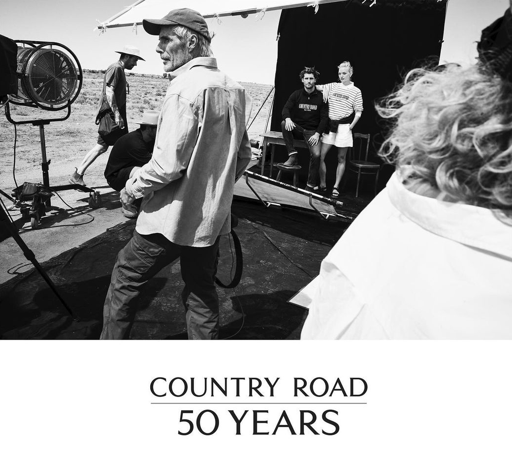 Black and white behind-the-scenes shot of a Country Road campaign, featuring crew, equipment, and models in a desert setting.