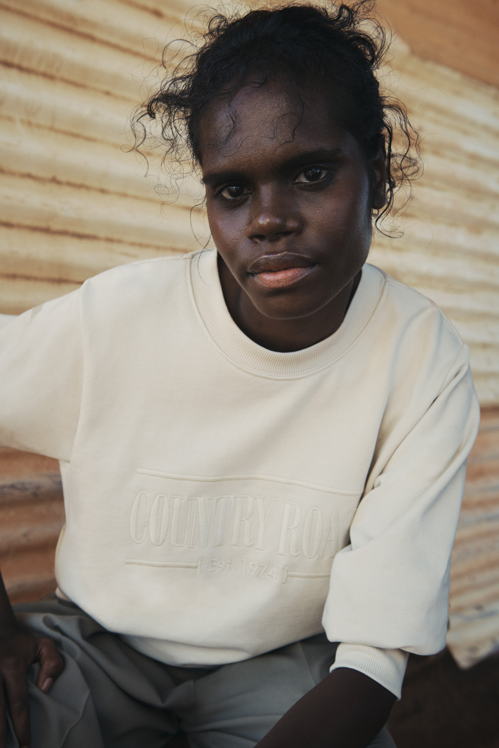 Close-up portrait of an Aboriginal woman in a beige sweatshirt looking directly at the camera with a neutral expression in an outdoor setting.