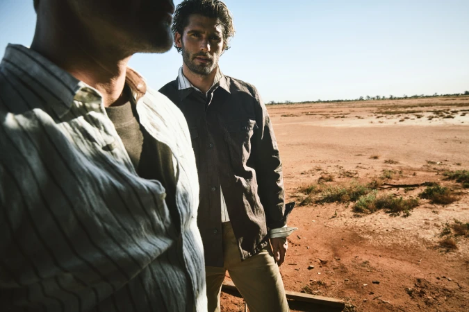 Two adult men stand in a sunlit, arid desert landscape, one in foreground and another in focus, wearing casual shirts and pants.