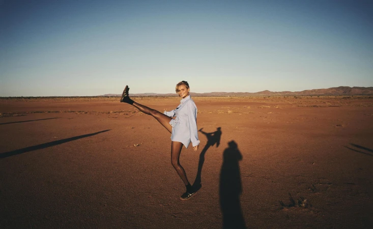 Woman in blue shirt and black shoes poses in desert with leg raised against blue sky, creating a playful and dynamic portrait.