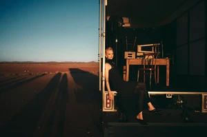 Blonde woman in a black dress seated on the back of a truck in a desert landscape during sunset for Country Road.