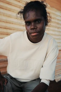 Portrait of an Indigenous woman wearing a cream sweatshirt with embroidered text against a corrugated iron background.