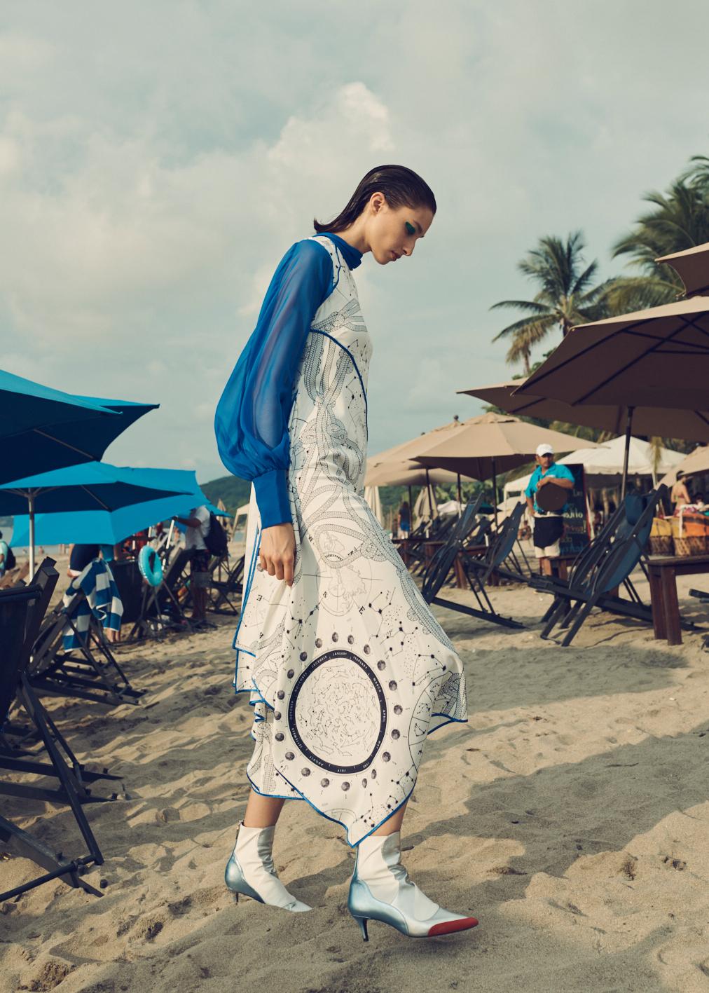 Fashion shot on a beach featuring a woman walking in a patterned dress with a blue blouse and light-colored ankle boots, ocean background.
