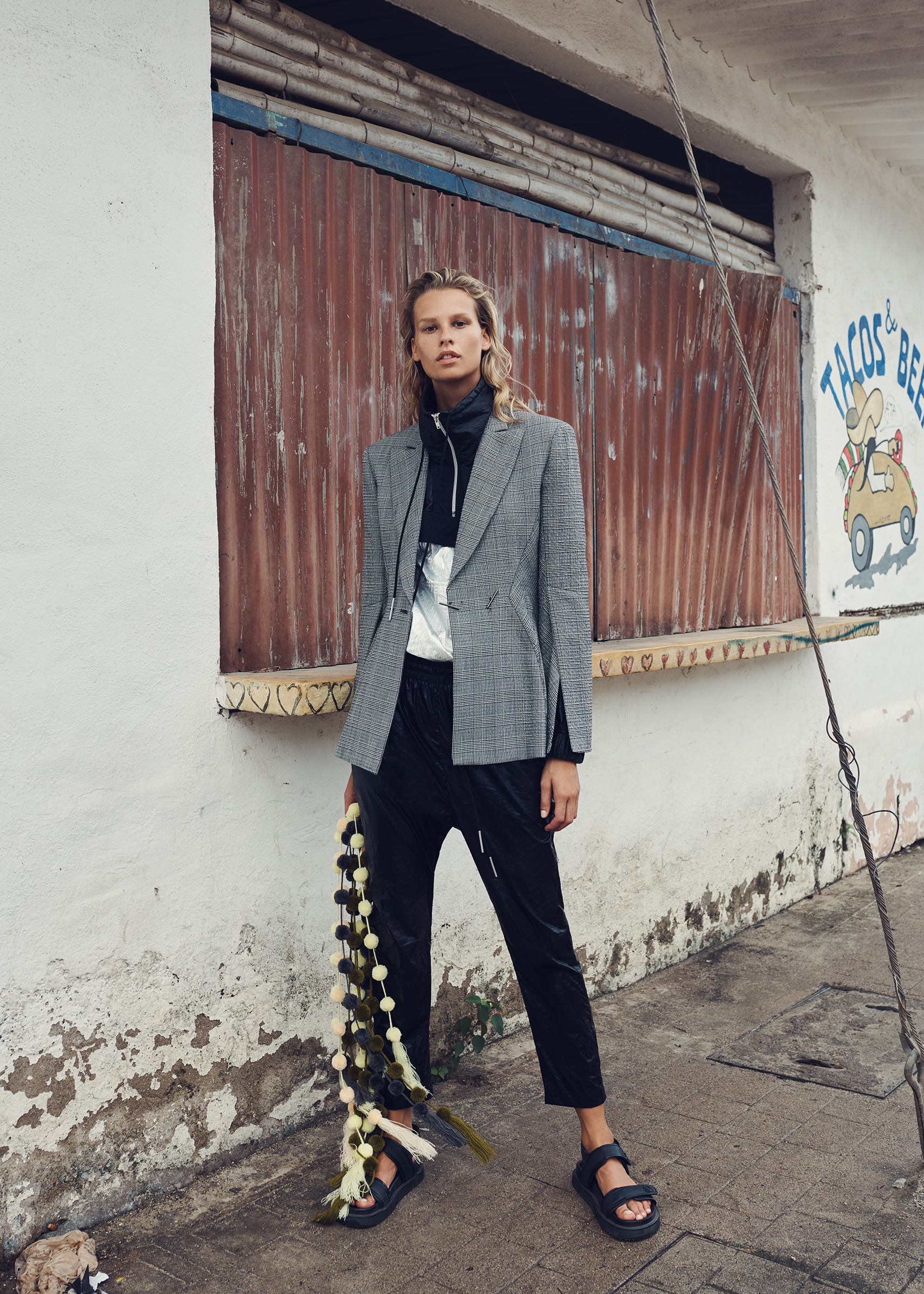 Fashionable woman in blazer and sandals poses on street by building with peeling paint and faded 'Tacos Beer' sign in daylight.
