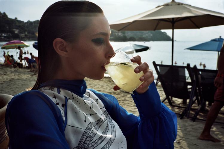 A woman drinks a cocktail while sitting on the beach in a close-up shot. The beach background includes umbrellas, chairs, and people.