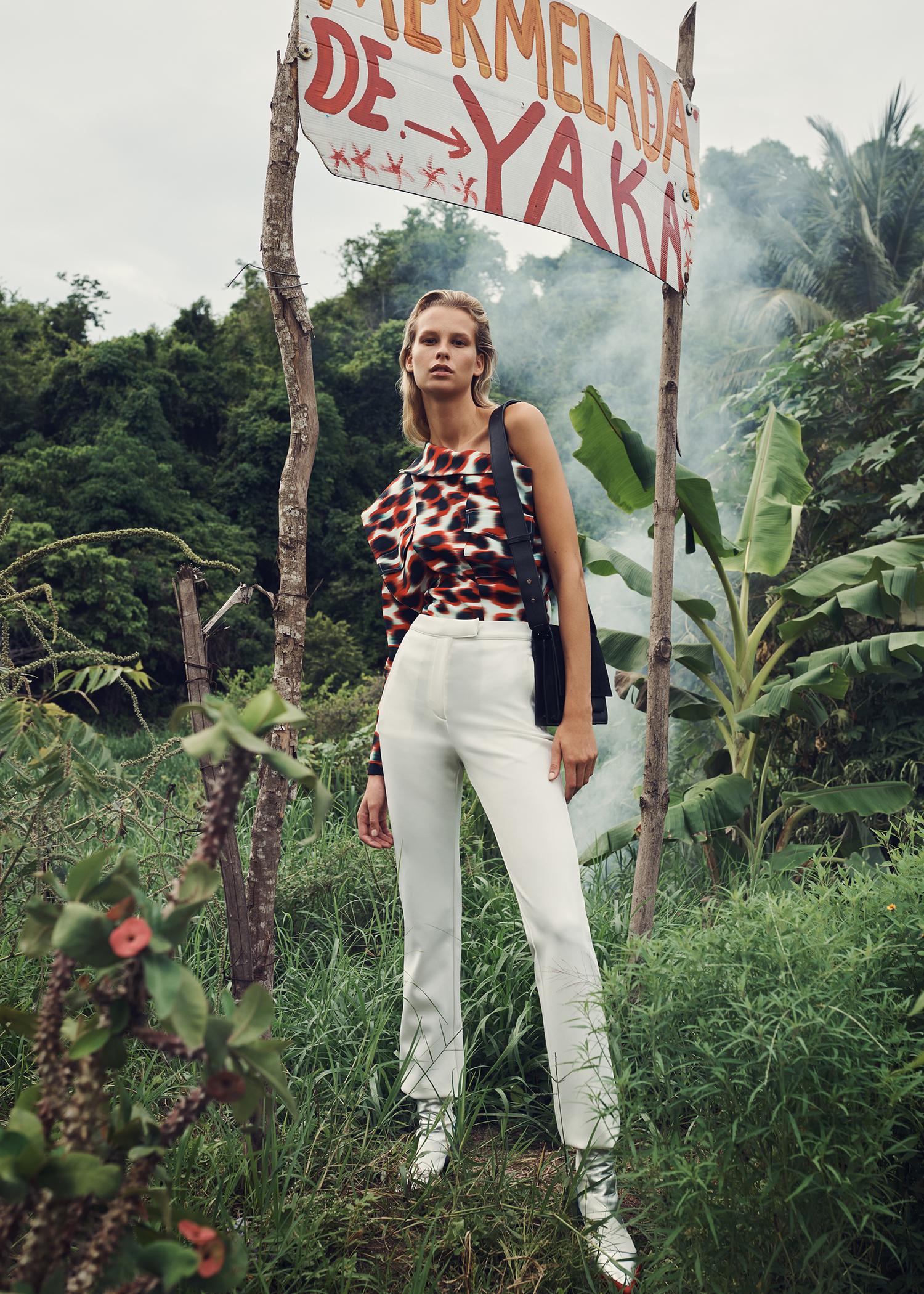 A blonde model in a brown patterned top and white pants stands next to a 'Mermelada de Yaka' sign in a lush jungle setting.