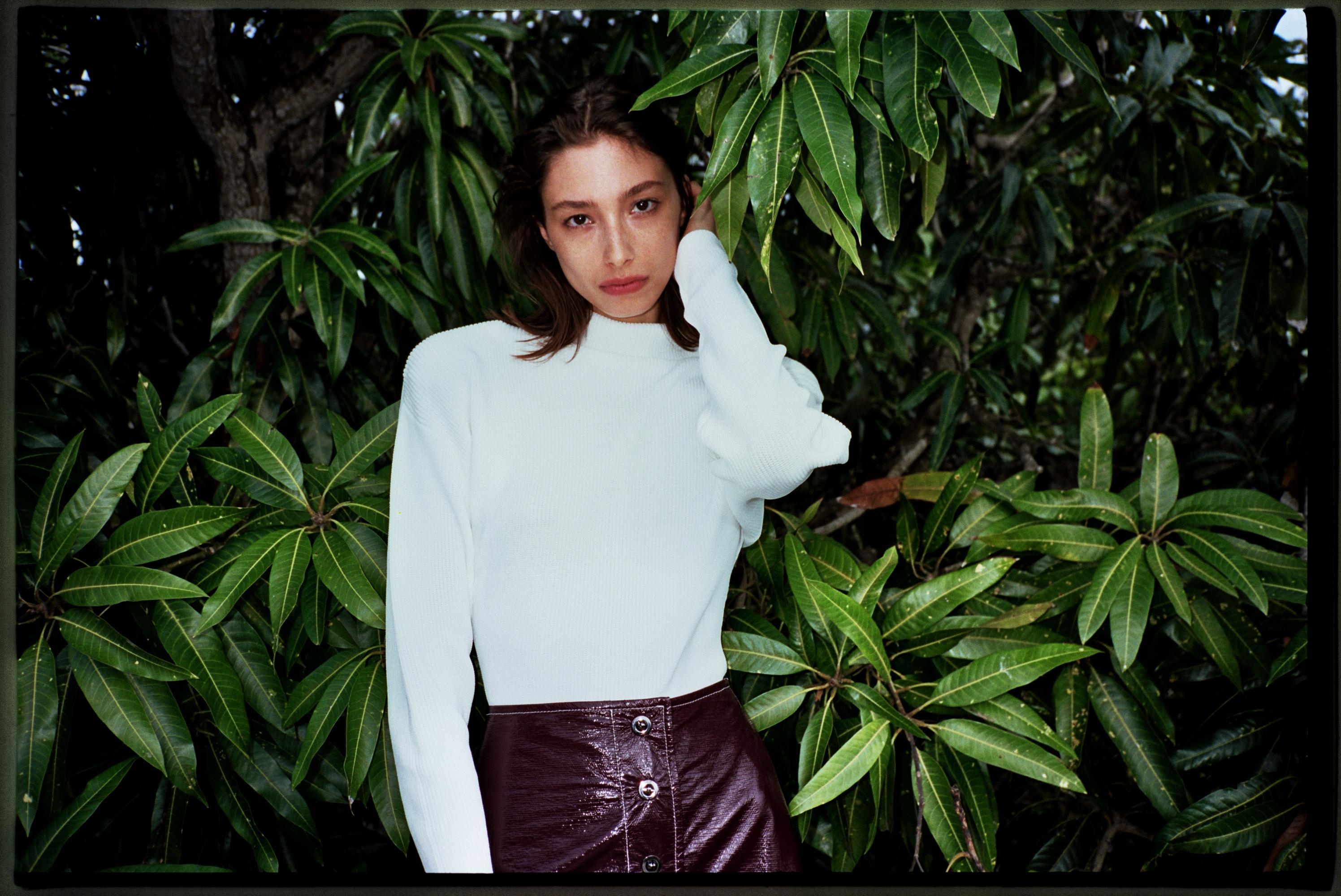 A young woman poses outdoors among lush green foliage, wearing a cream long-sleeve top and a short, dark-burgundy patent leather skirt.