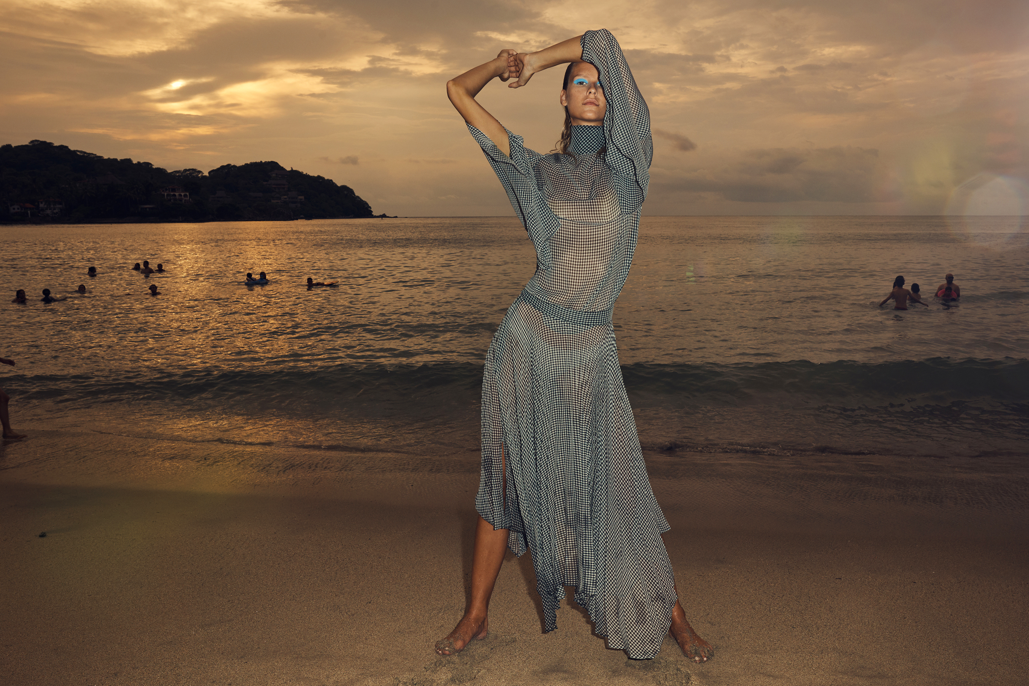 Fashion shot of a woman in a sheer patterned dress posing on a beach at sunset, with ocean swimmers visible in the background.