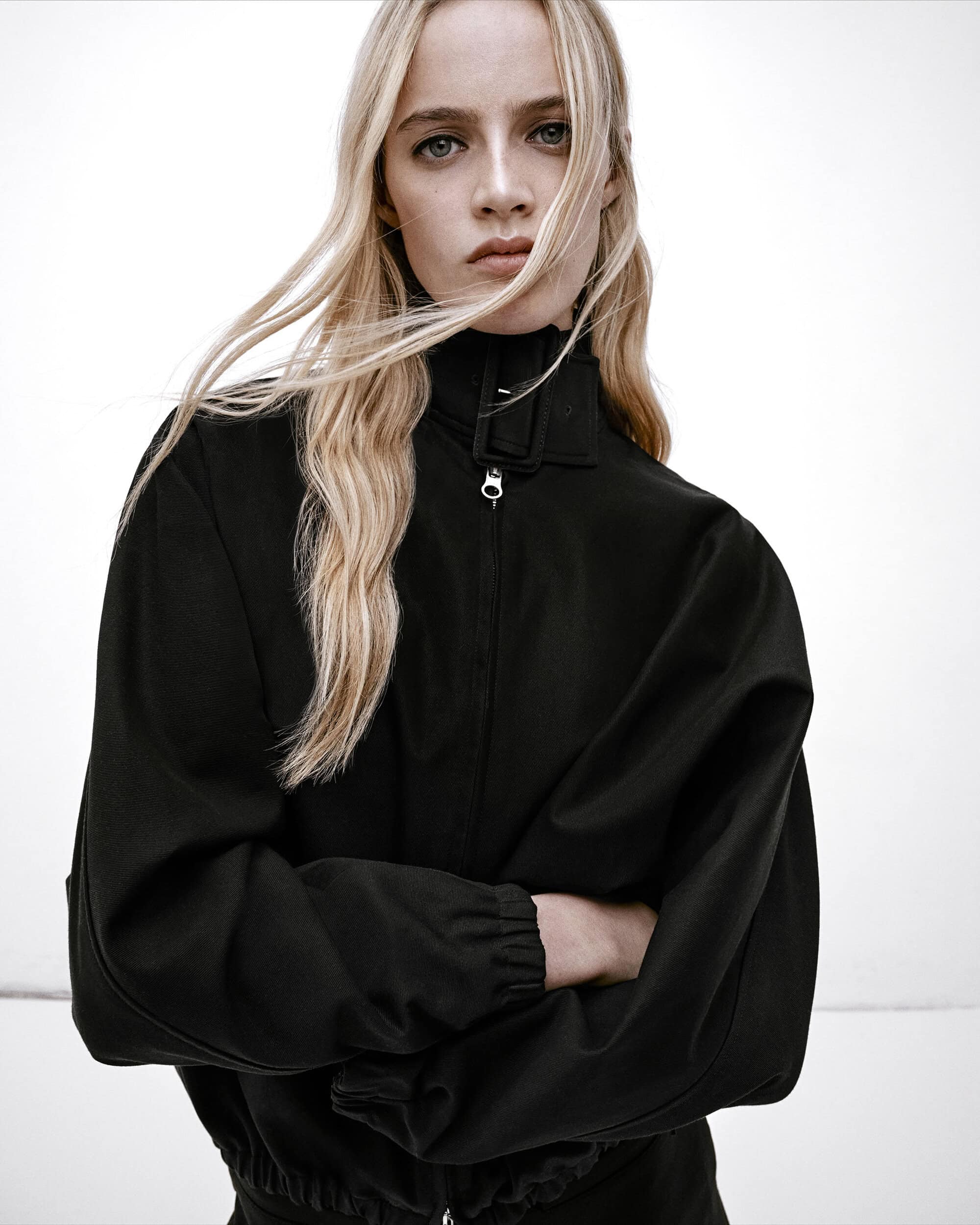 Model with long blonde hair wearing a black high-collar jacket against a plain white background