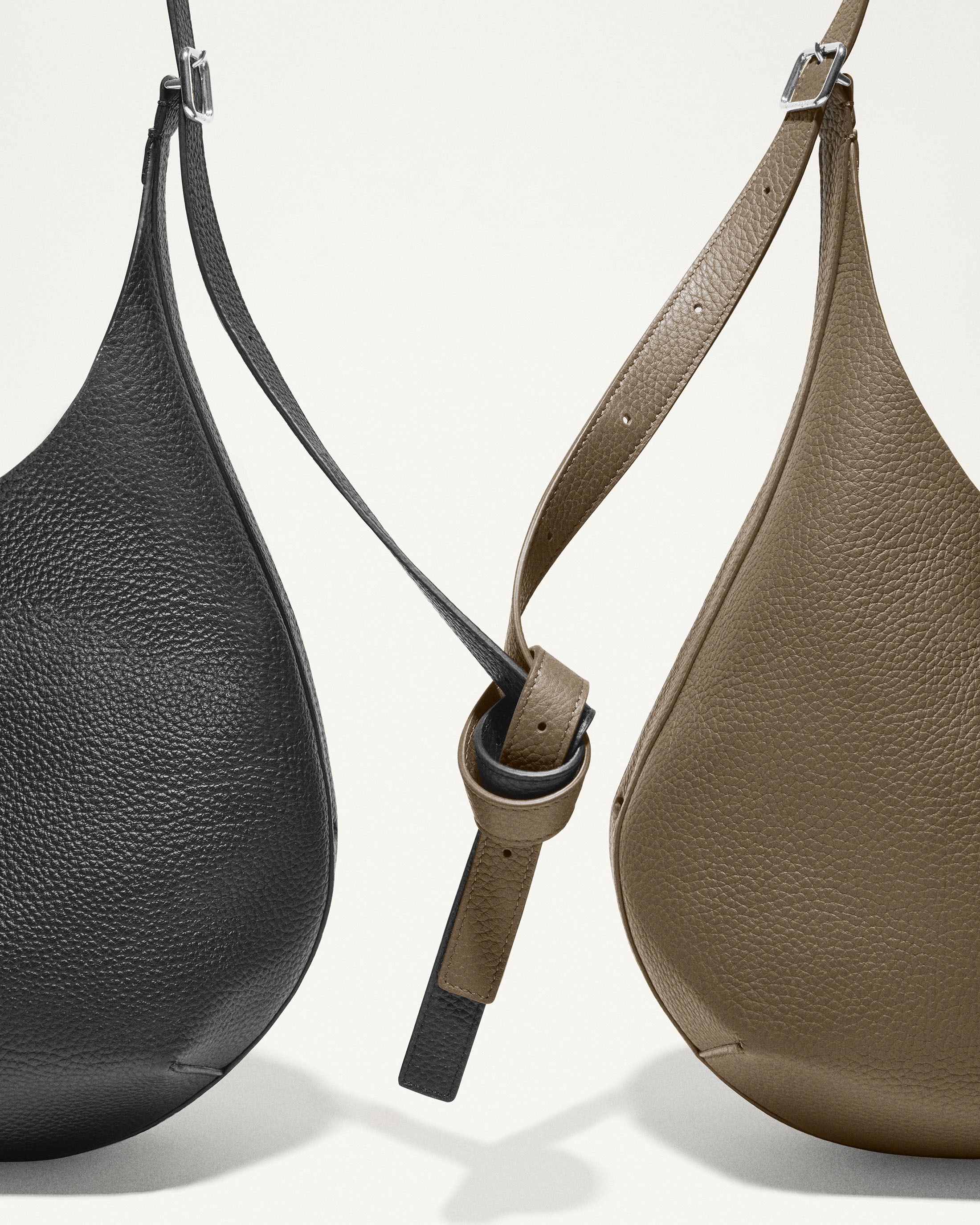 Studio shot of two luxury shoulder bags in black and taupe. Bags have a grainy leather texture with adjustable straps and silver hardware.