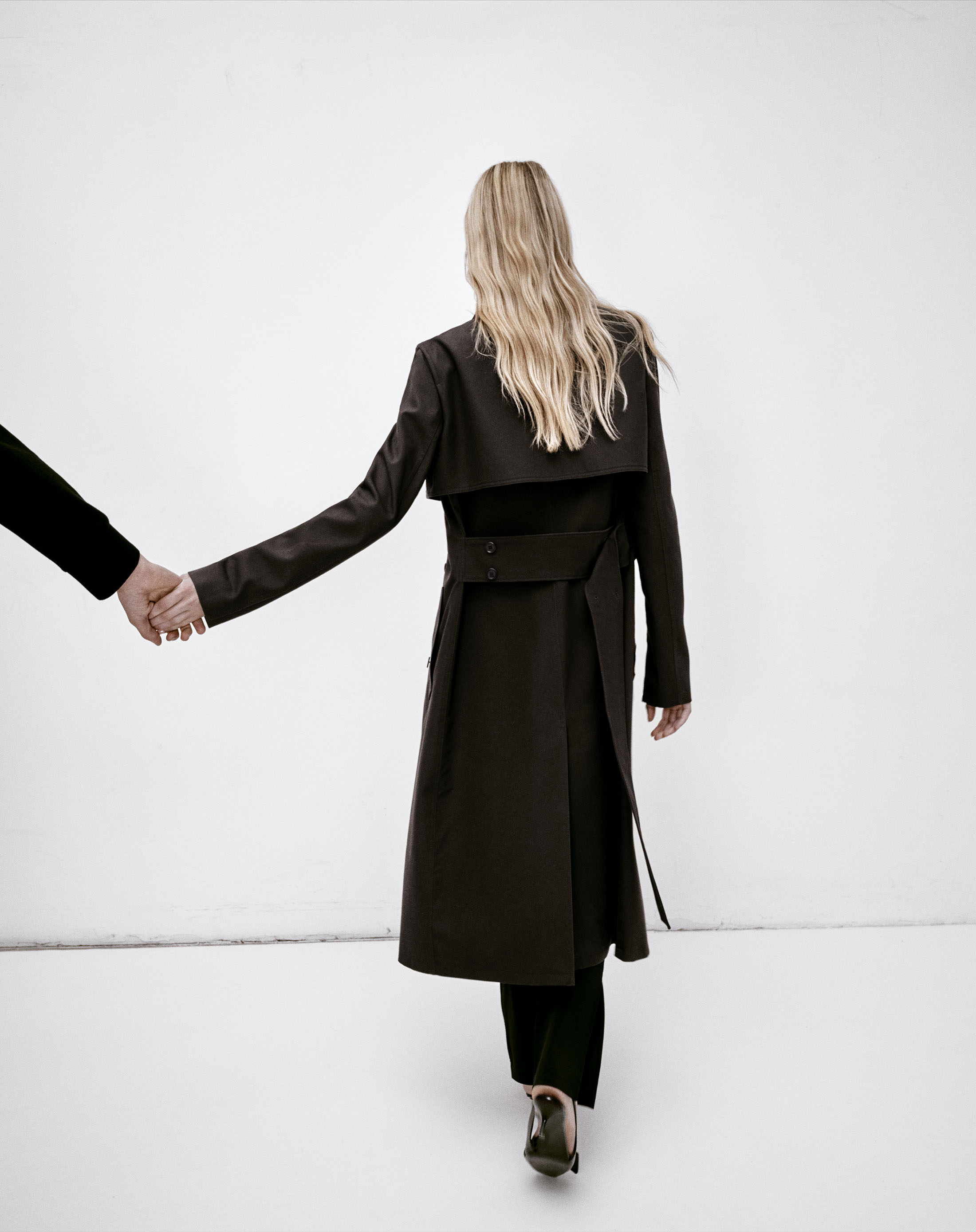 Model with long blonde hair seen from behind wearing a dark brown coat holding hands with another person