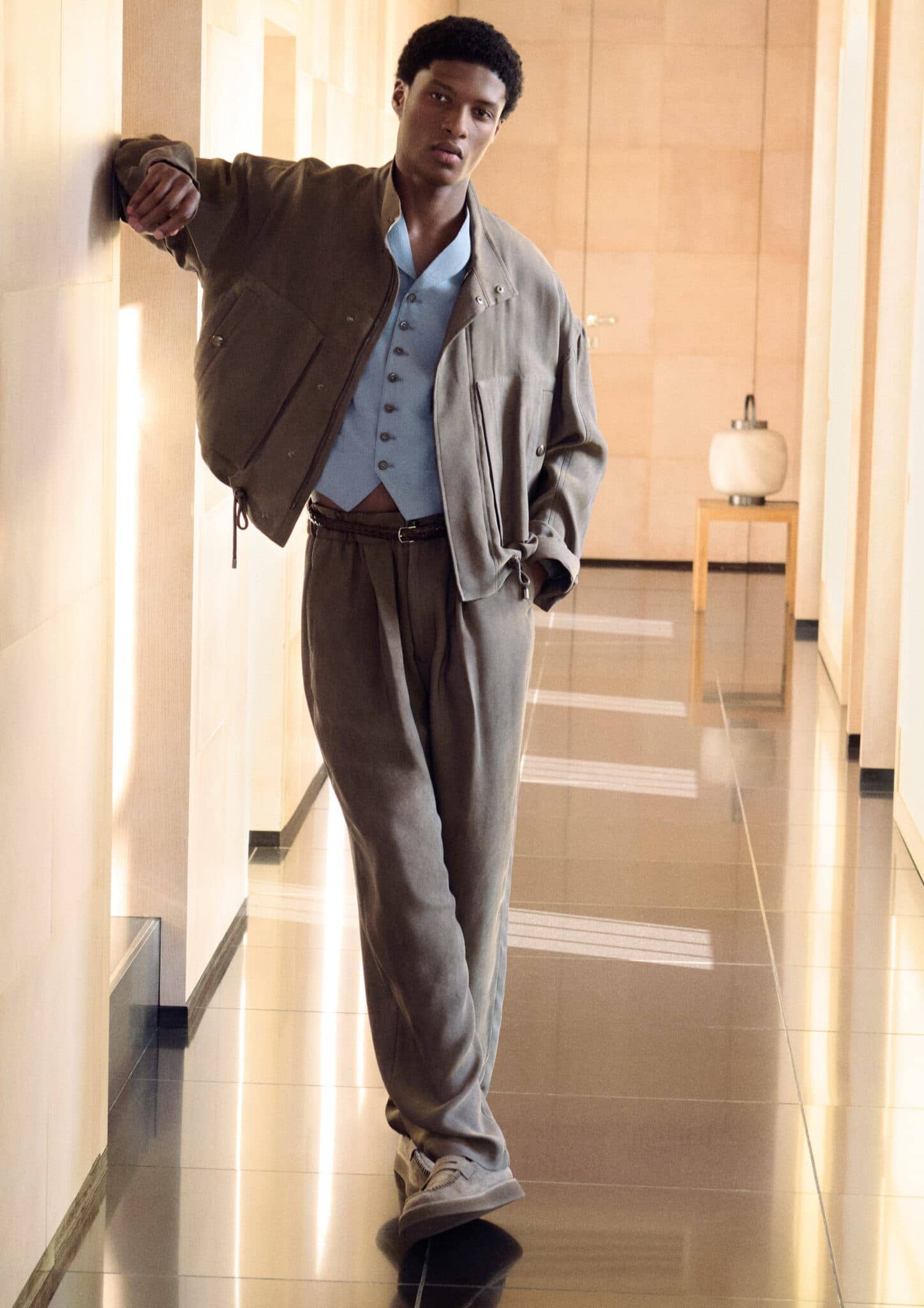 Dark-skinned man in vest, jacket, trousers posing indoors. Soft, diffused lighting shows neutral tones and modern architectural details.