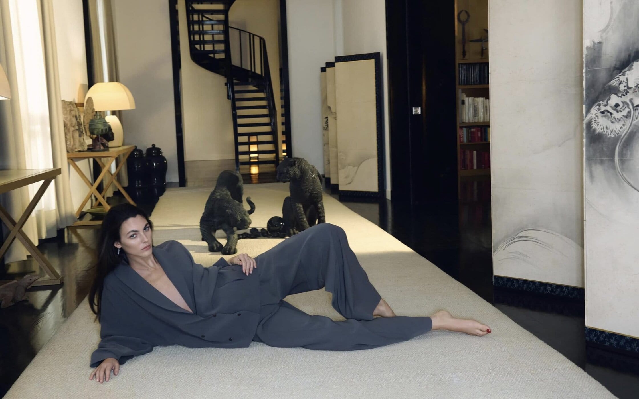 A woman poses in a grey suit on a light carpet in a modern room with animal sculptures, lamps, a black spiral staircase, and artwork.
