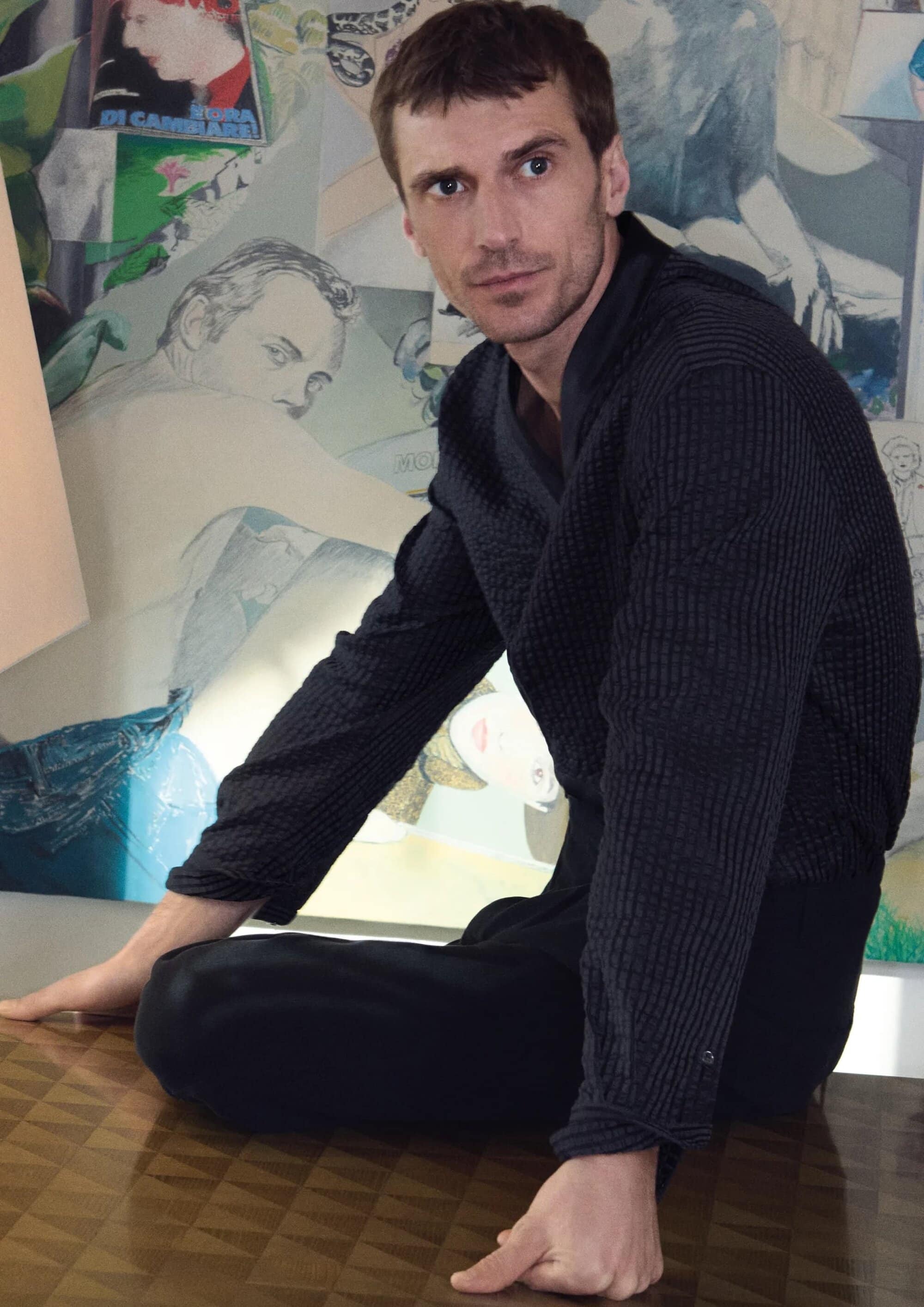 Adult male sits on a reflective surface with a collage-style art background. He is wearing a dark textured shirt and dark pants.