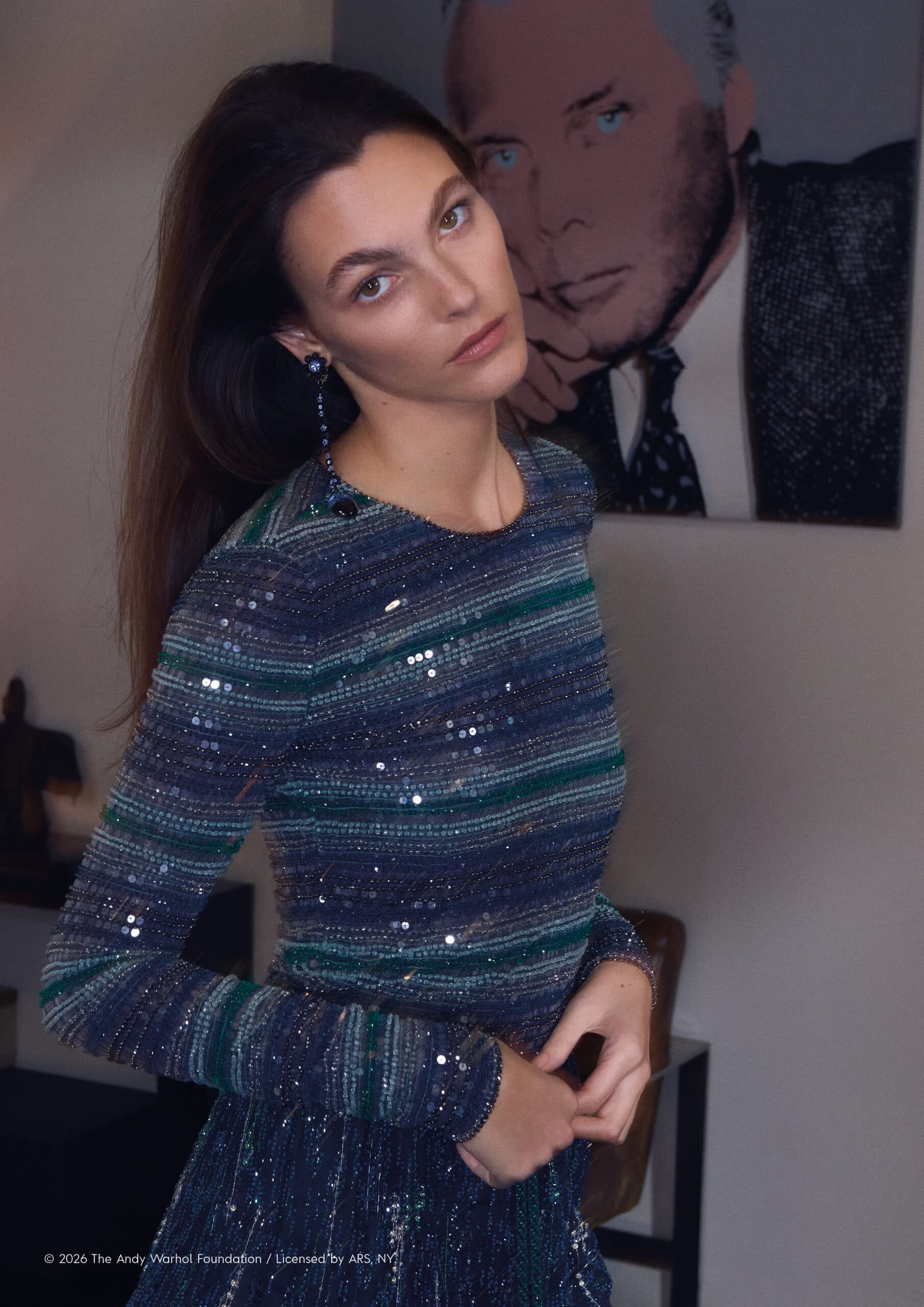 Portrait of a woman in a sequined dress posing indoors with neutral wall, a framed portrait, and minimal furniture in the background.