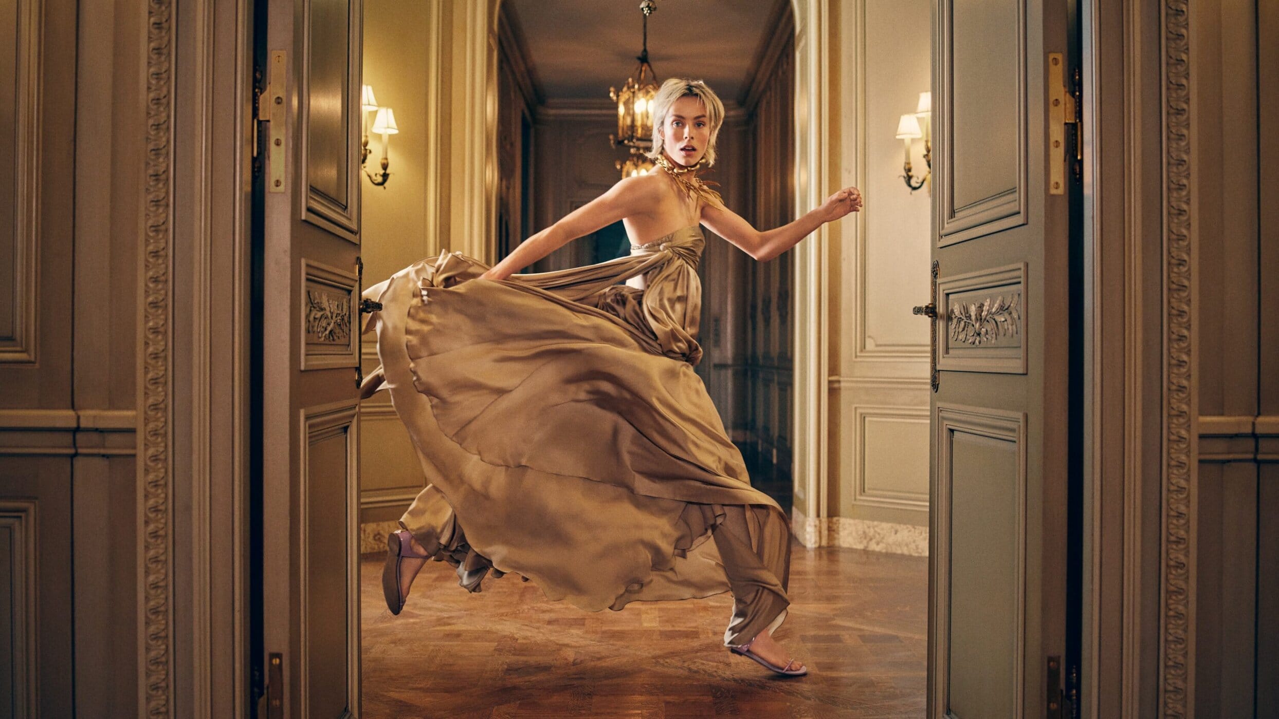 A woman in a gold dress runs through an open doorway inside an ornate room with hardwood floors and neutral-colored walls and trim.