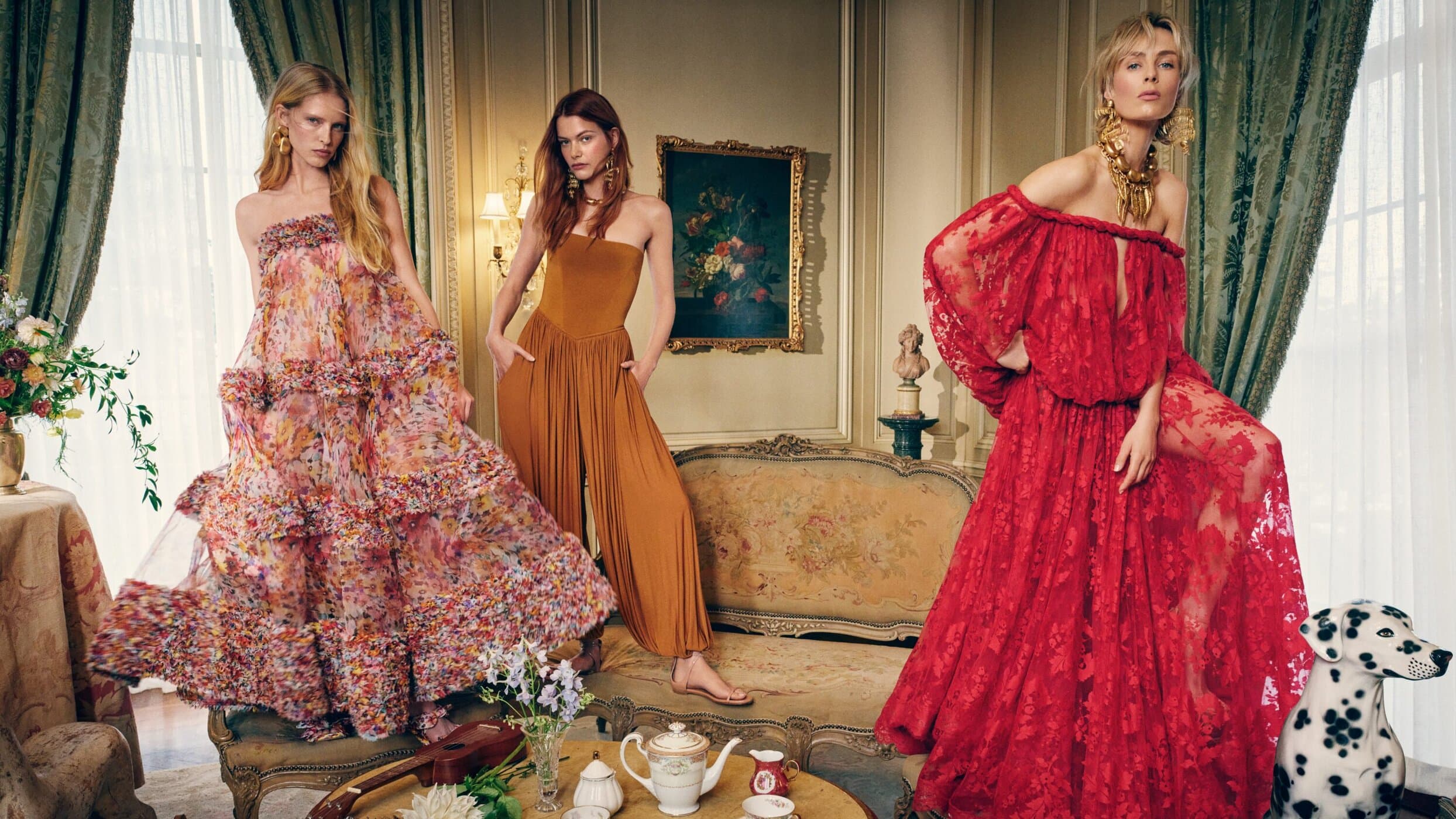 Three fashion models pose indoors, showcasing ornate red and floral gowns, accented by statement jewelry and detailed interior decor.