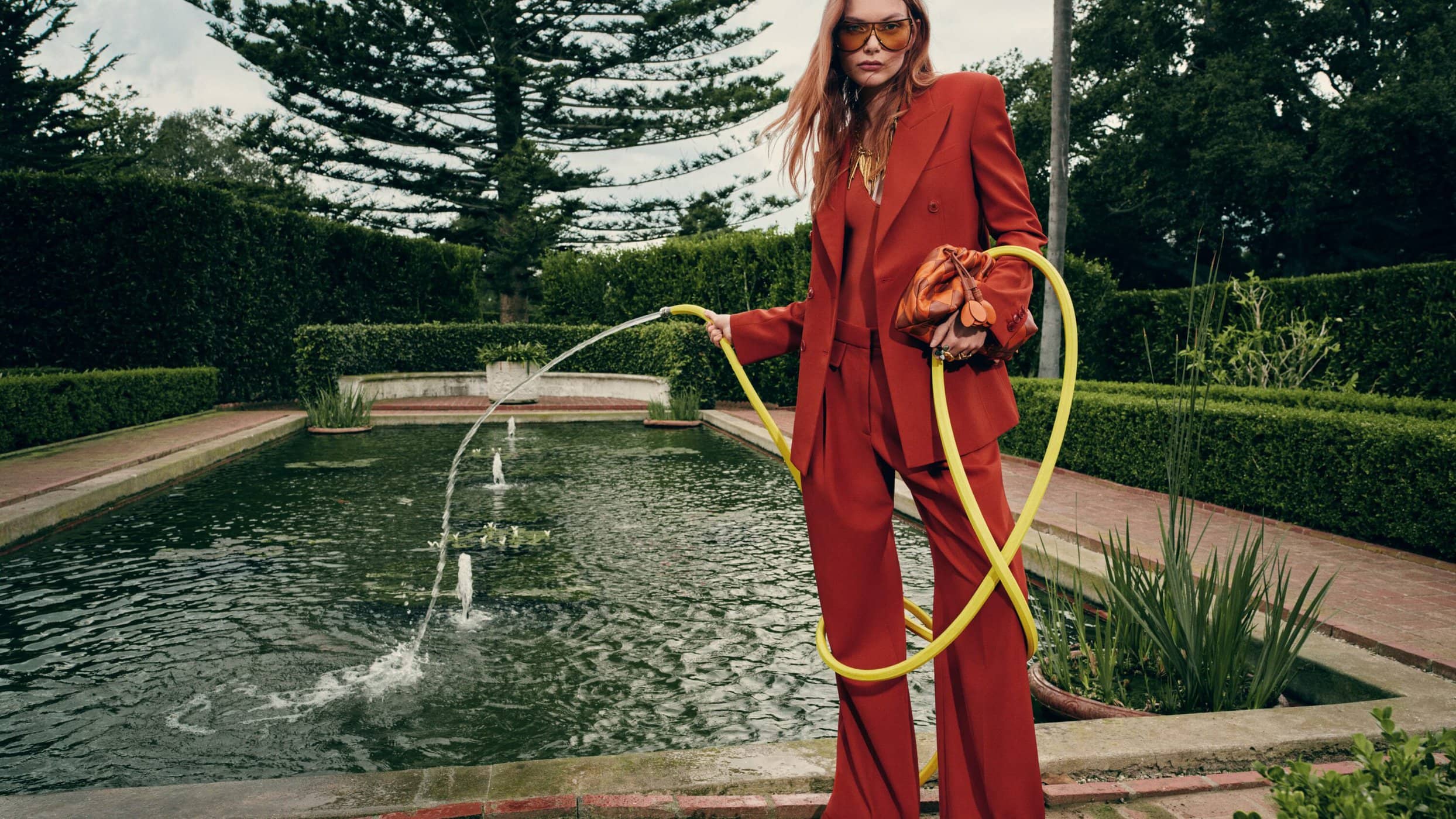 A woman in a red suit stands beside a pond, holding a yellow hose, wearing sunglasses, and carrying a bag in an outdoor fashion shoot.