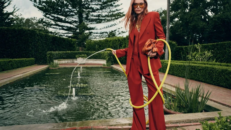 A woman in a red suit stands beside a pond, holding a yellow hose, wearing sunglasses, and carrying a bag in an outdoor fashion shoot.