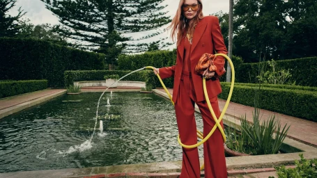 Model in a red pantsuit holding a yellow garden hose by a reflecting pool for Zimmermann Spring 2026 Ad Campaign