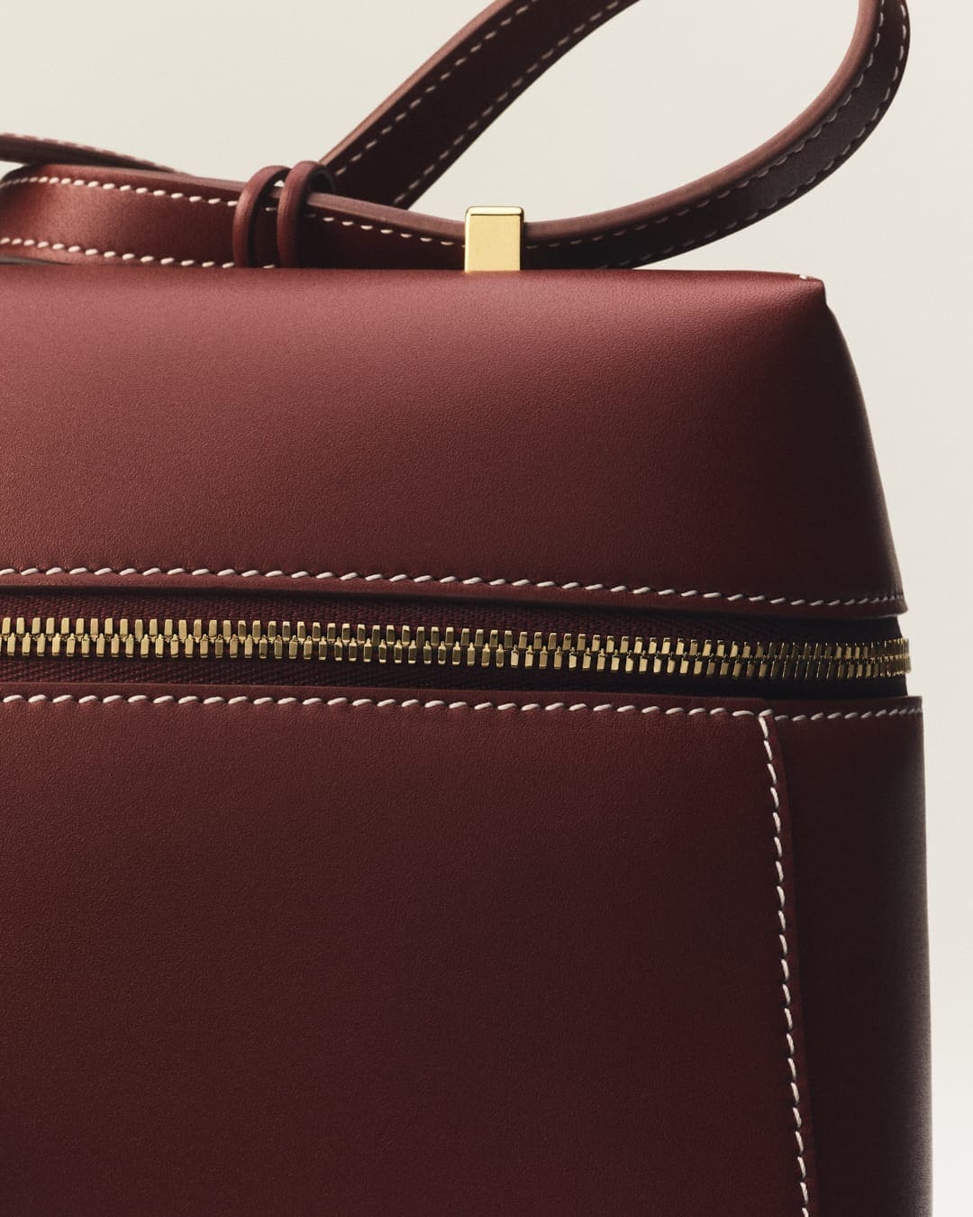 Close-up of a burgundy leather handbag with gold hardware and visible stitching, set against a white background in a studio setting.