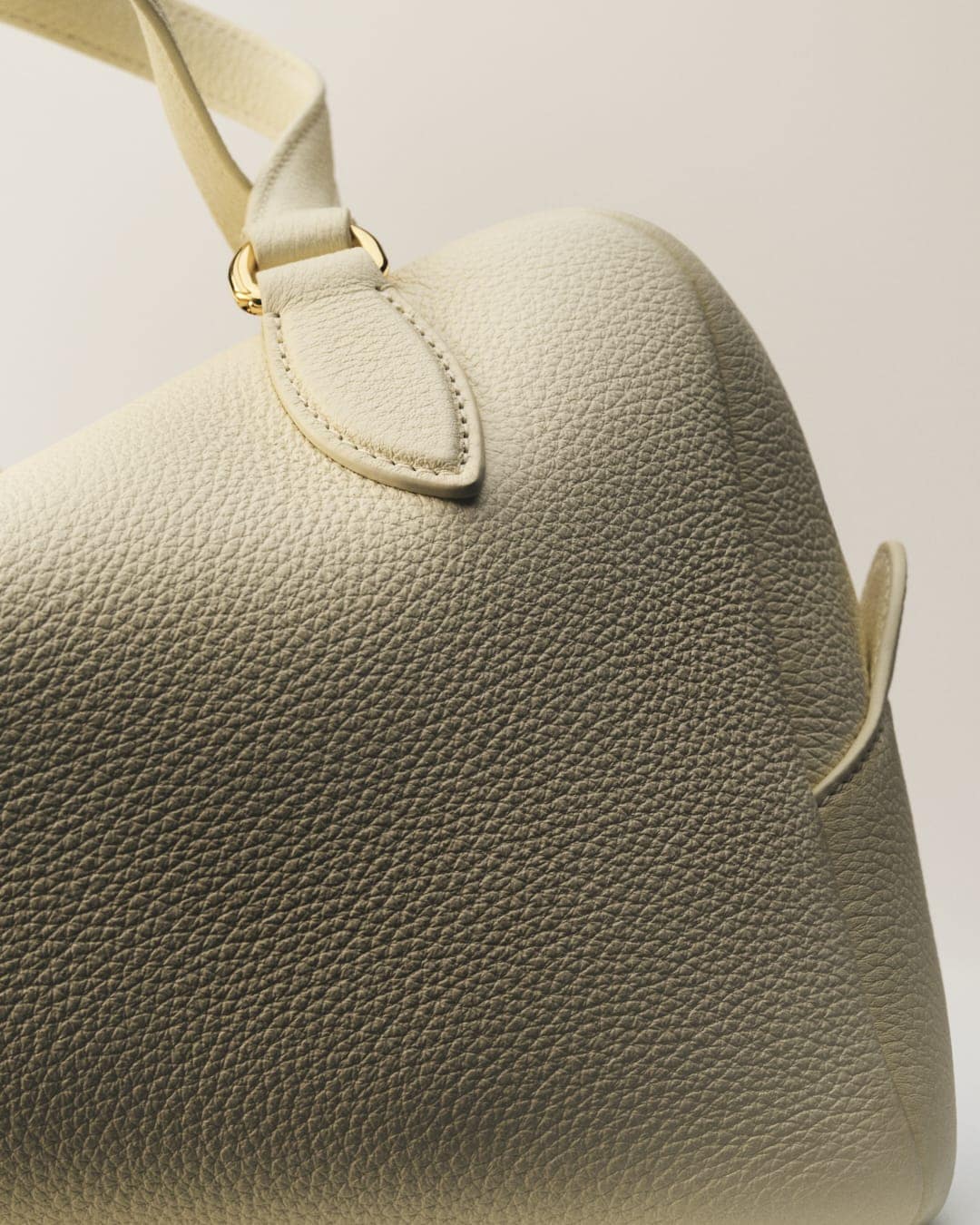 Close-up of a textured, off-white leather handbag with gold hardware and soft lighting in a studio setting; high-fashion aesthetic.