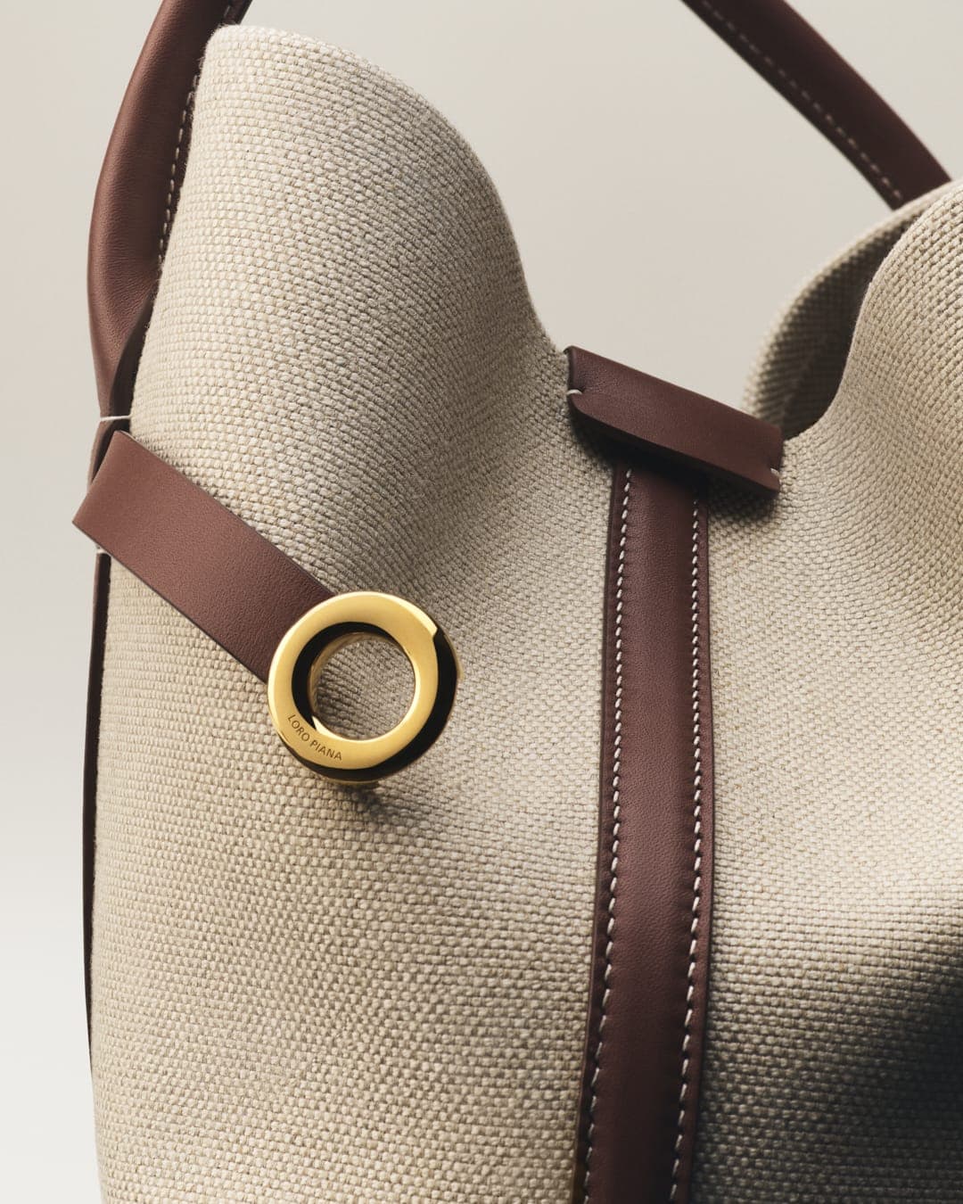Tote bag with brown leather straps and gold hardware. Neutral color canvas material. Fashionable everyday carryall accessory.