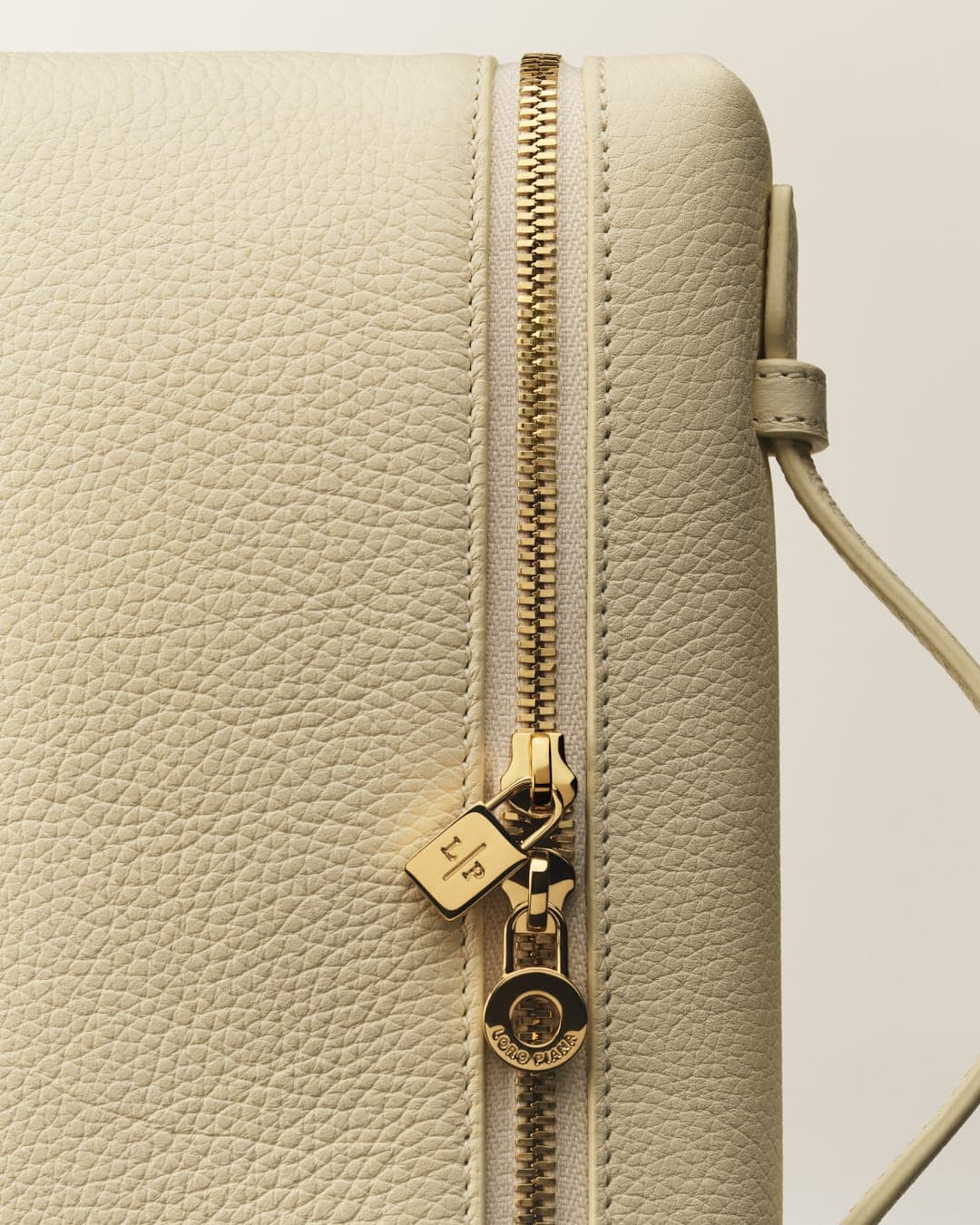 Close-up on a cream-colored leather bag with a gold zipper, featuring a branded pull tab, set against a light, neutral background.