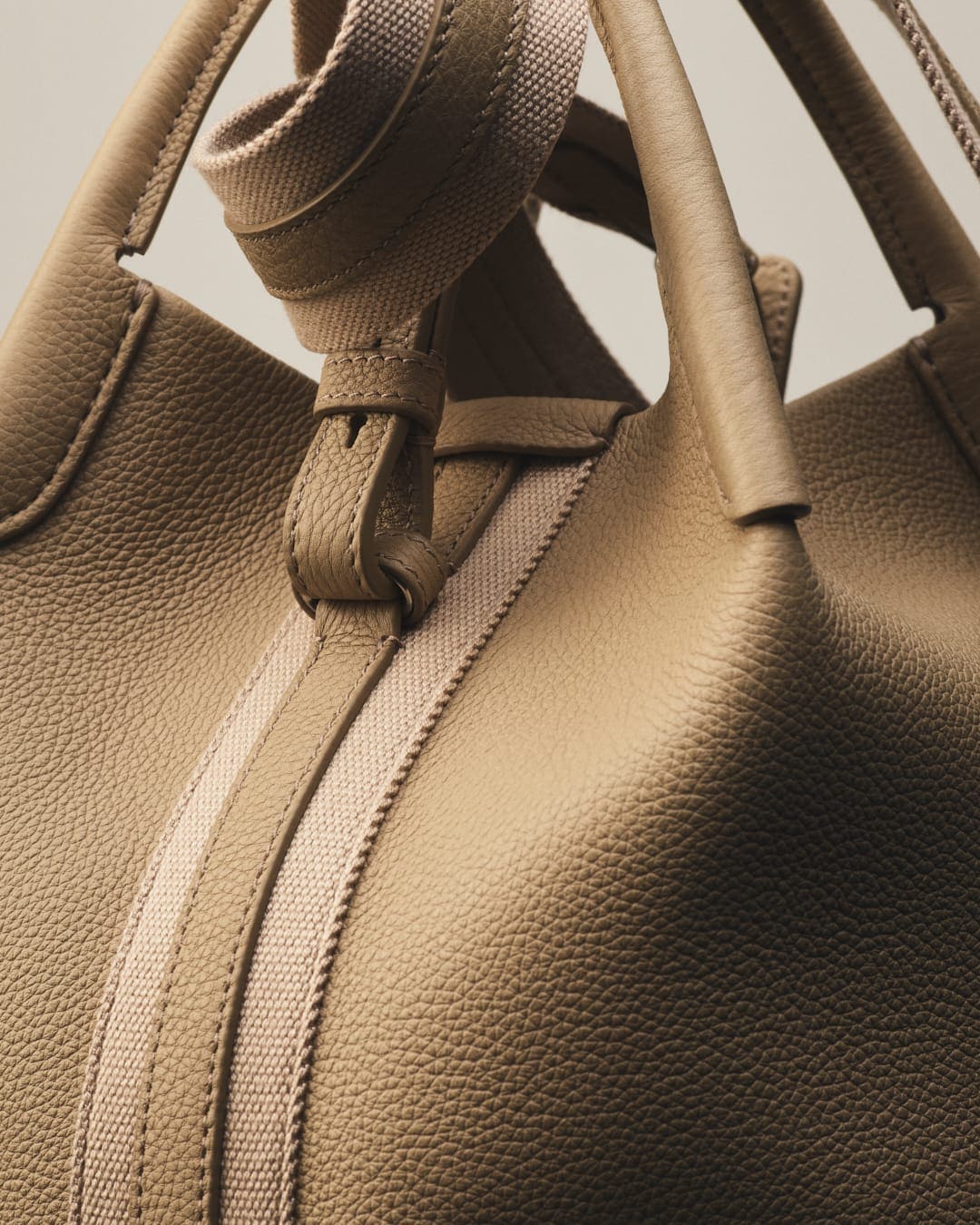 Close-up of a tan leather tote bag with rolled handles and a beige woven strap, set against a soft, neutral background.
