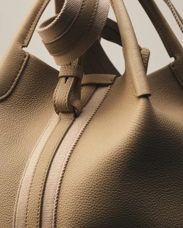 Close-up of a tan leather tote bag with rolled handles and a beige woven strap, set against a soft, neutral background.