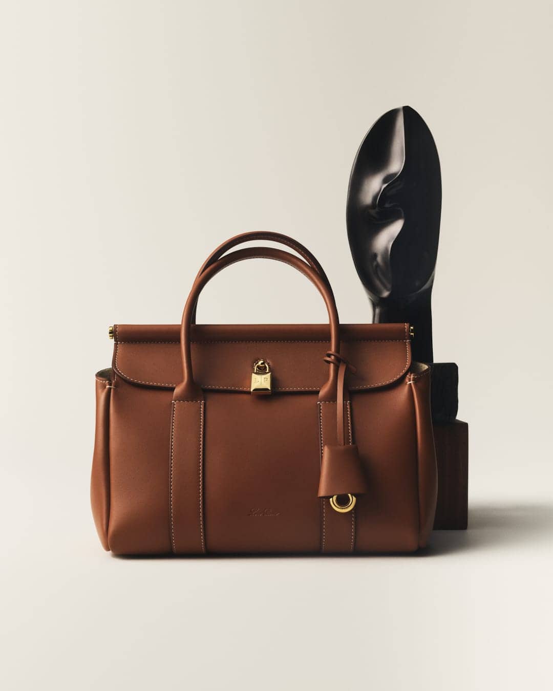 Elevated studio shot of a brown leather satchel handbag with gold hardware, standing next to a glossy black sculpture.