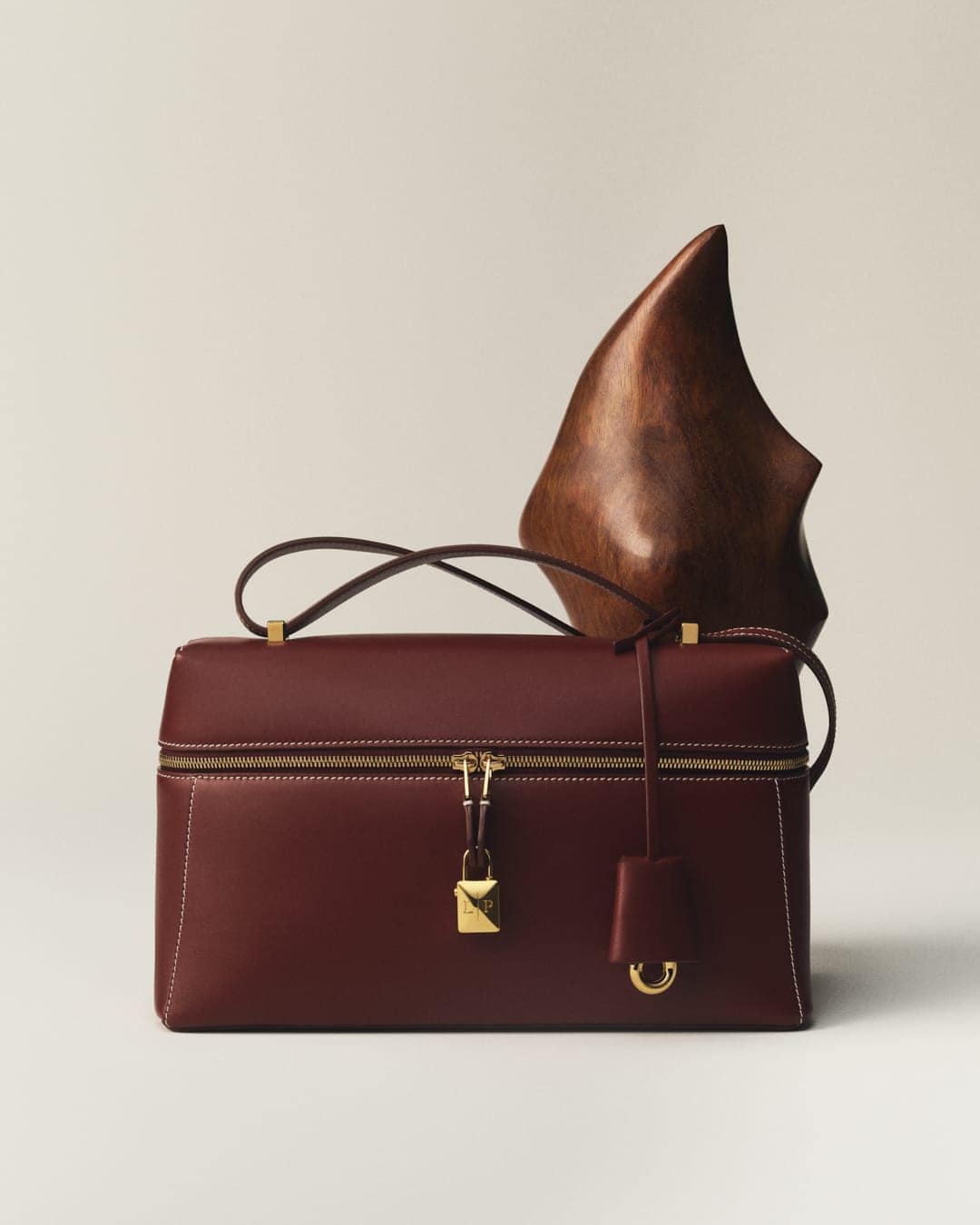 Burgundy leather box handbag with gold hardware and a textured brown sculpture on a light background, product shot for fashion.