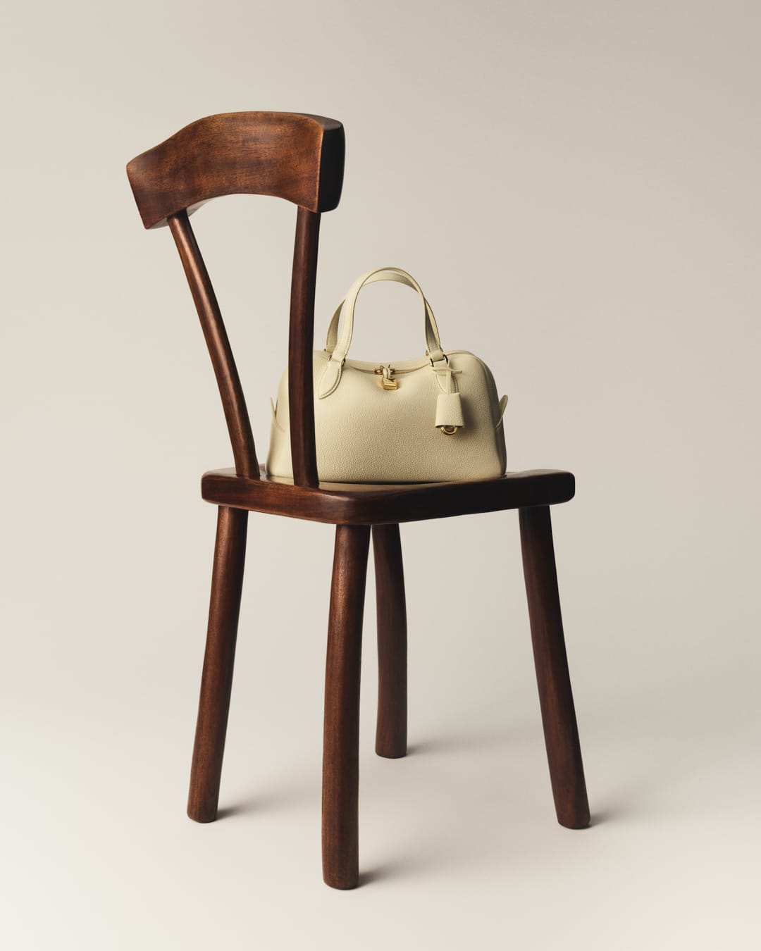 Ivory-colored leather handbag with top handles sitting on a dark wood chair against a soft neutral background in a studio setting.