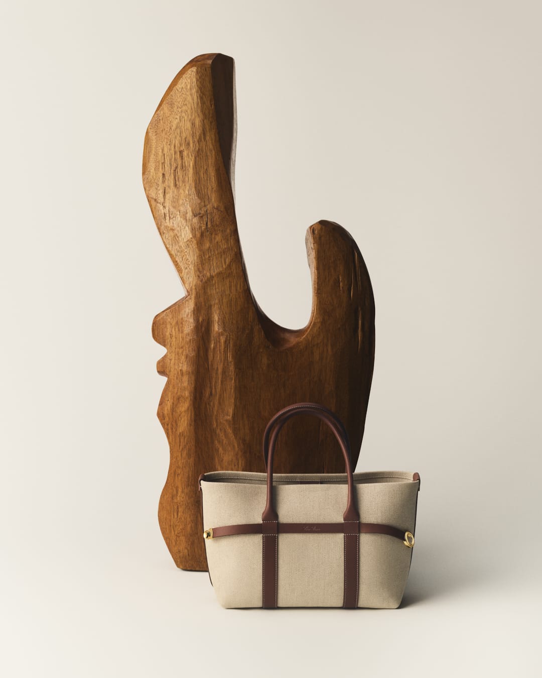 Elegant beige tote bag with brown leather accents showcased alongside a carved wooden sculpture against a neutral backdrop.