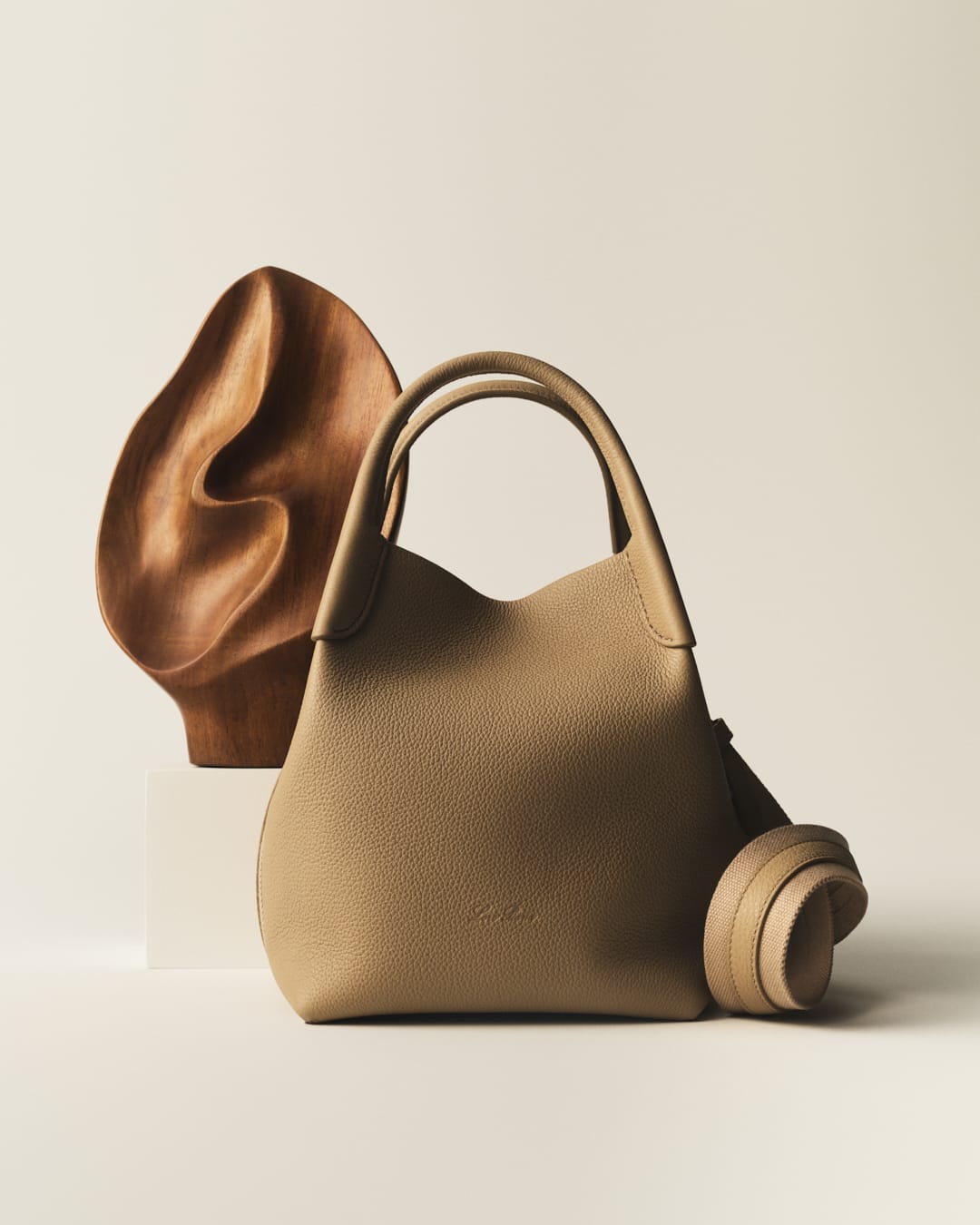 Tan leather tote bag with shoulder strap and wooden sculpture, studio shot on neutral background, product photography for fashion accessory.