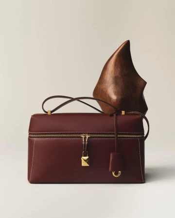A burgundy leather handbag with gold hardware next to a carved wooden sculpture on a neutral white backdrop.