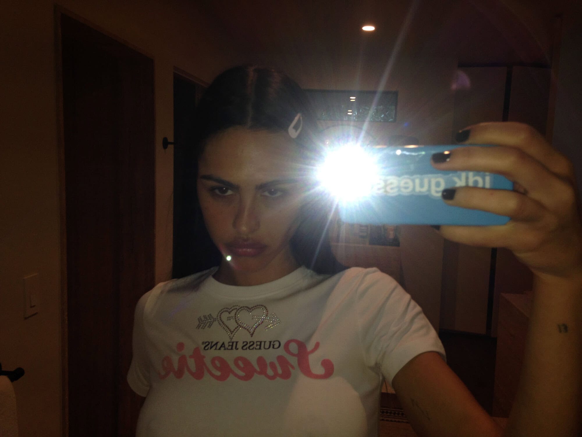 Brunette woman with dark fingernails and a white T-shirt takes a mirror selfie indoors with a phone flash creating a lens flare.