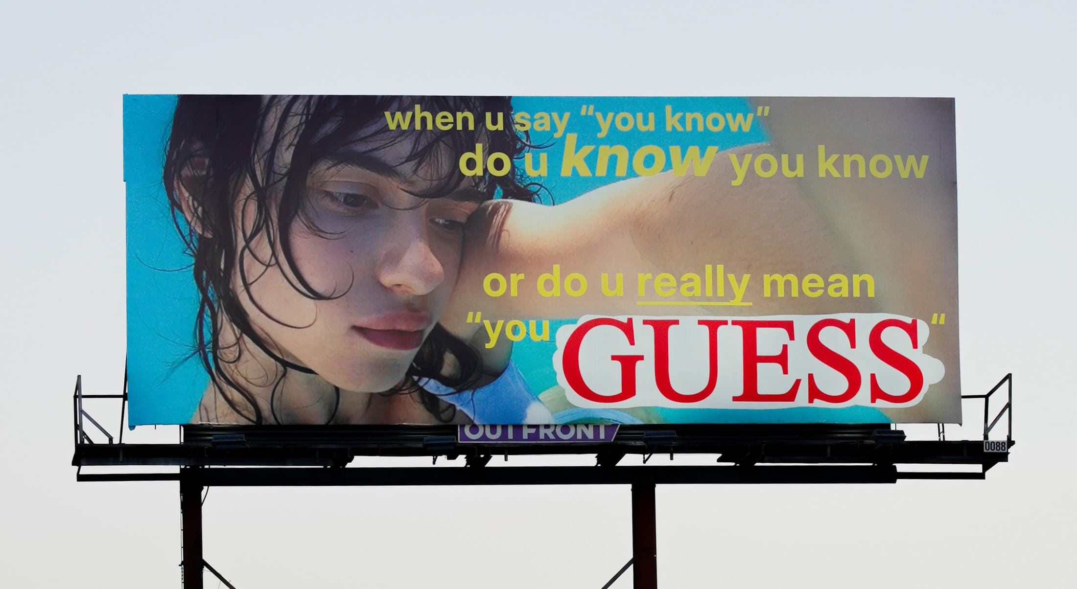 Billboard advertising Guess features a wet-haired model against a blue background, with overlaid yellow and red text, against a blue sky.