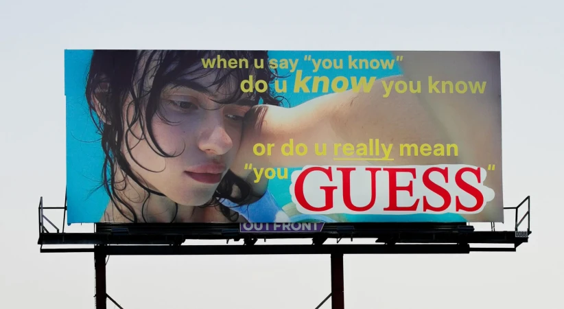 Billboard advertising Guess features a wet-haired model against a blue background, with overlaid yellow and red text, against a blue sky.