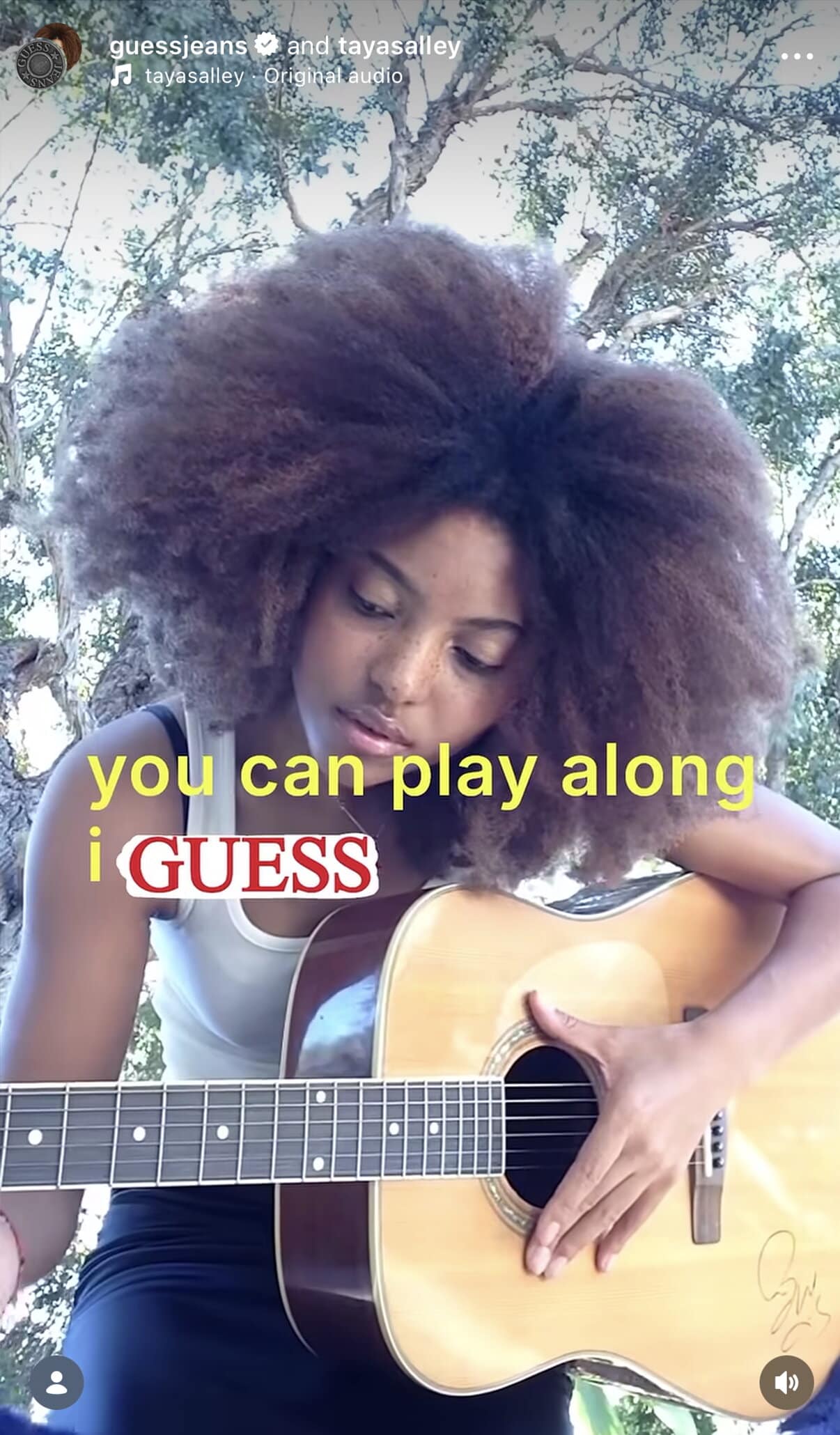 Woman with large afro plays an acoustic guitar outdoors, wearing a black outfit with a light-colored guitar, surrounded by trees in daylight.