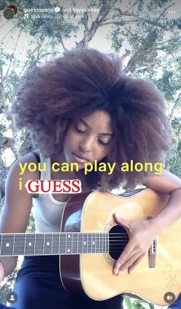 Woman with large afro plays an acoustic guitar outdoors, wearing a black outfit with a light-colored guitar, surrounded by trees in daylight.