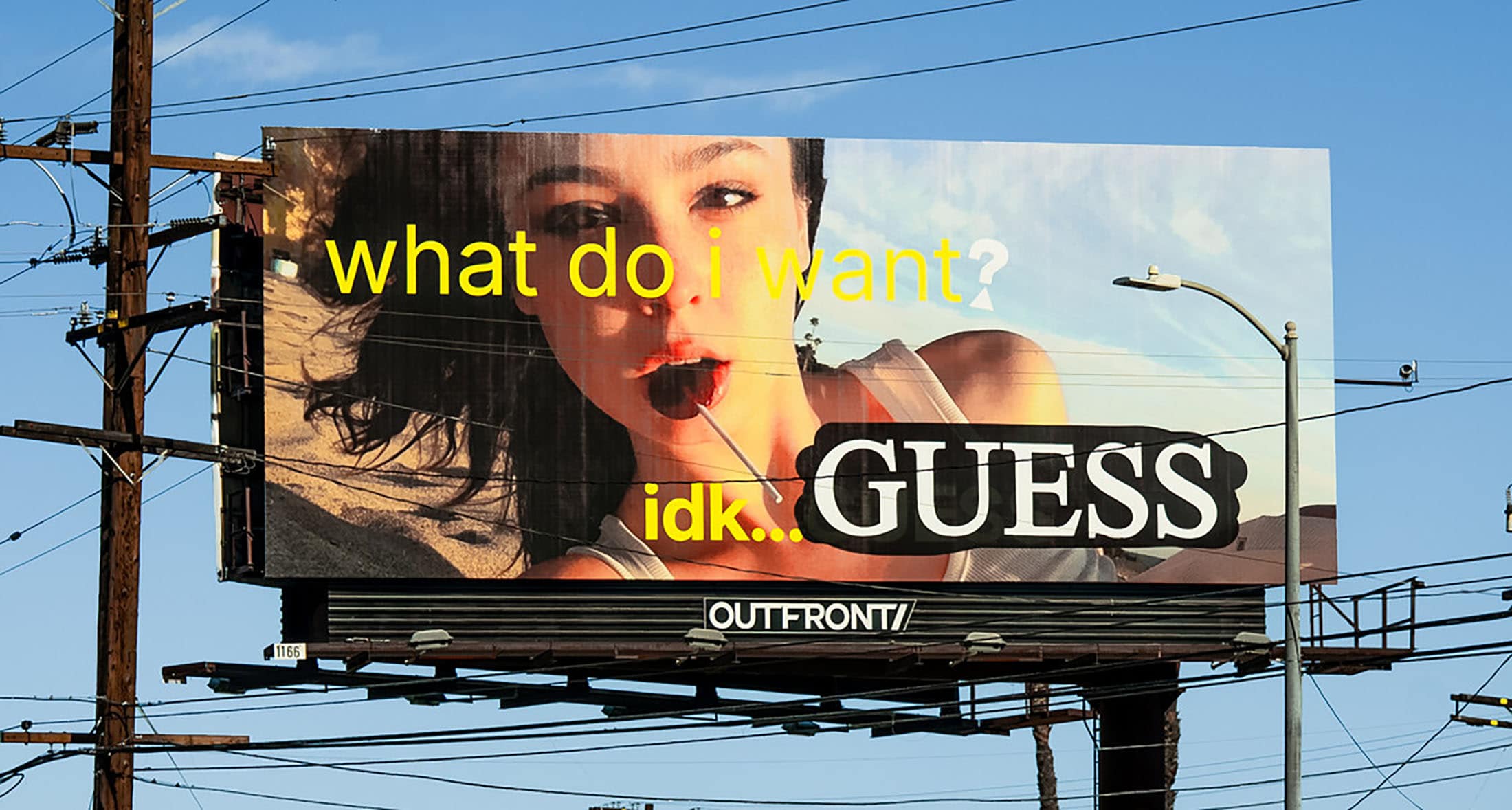 Eye-level billboard featuring a woman with an open mouth and cherry lollipop, next to a Guess logo, against a partly cloudy blue sky.
