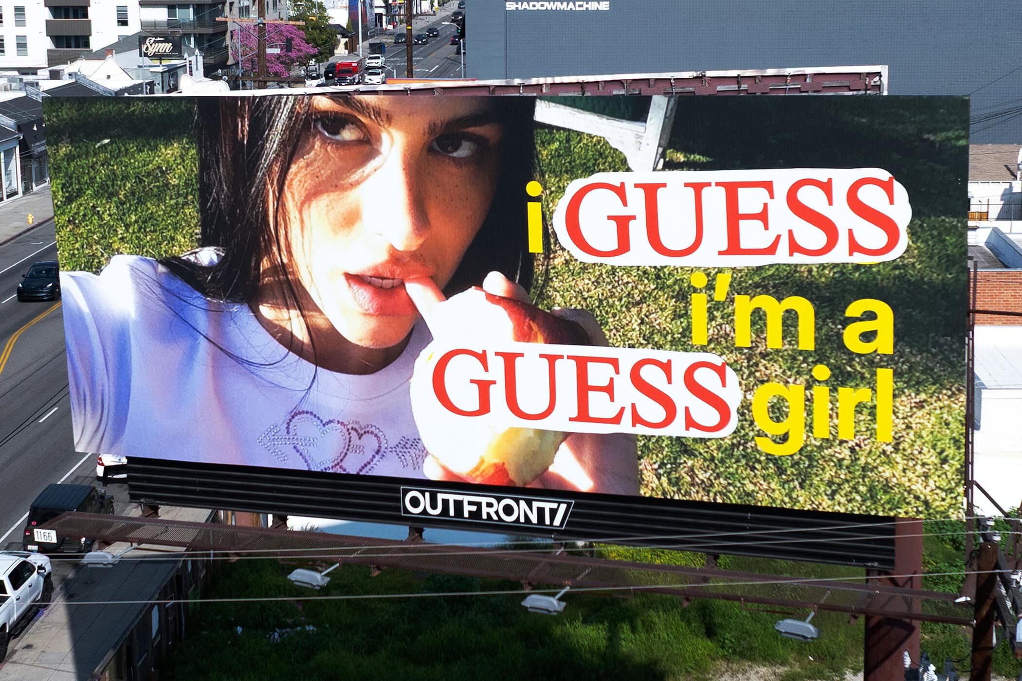 Billboard featuring a model promoting Guess, with green grass background and the Guess logo prominently displayed, captured outdoors.