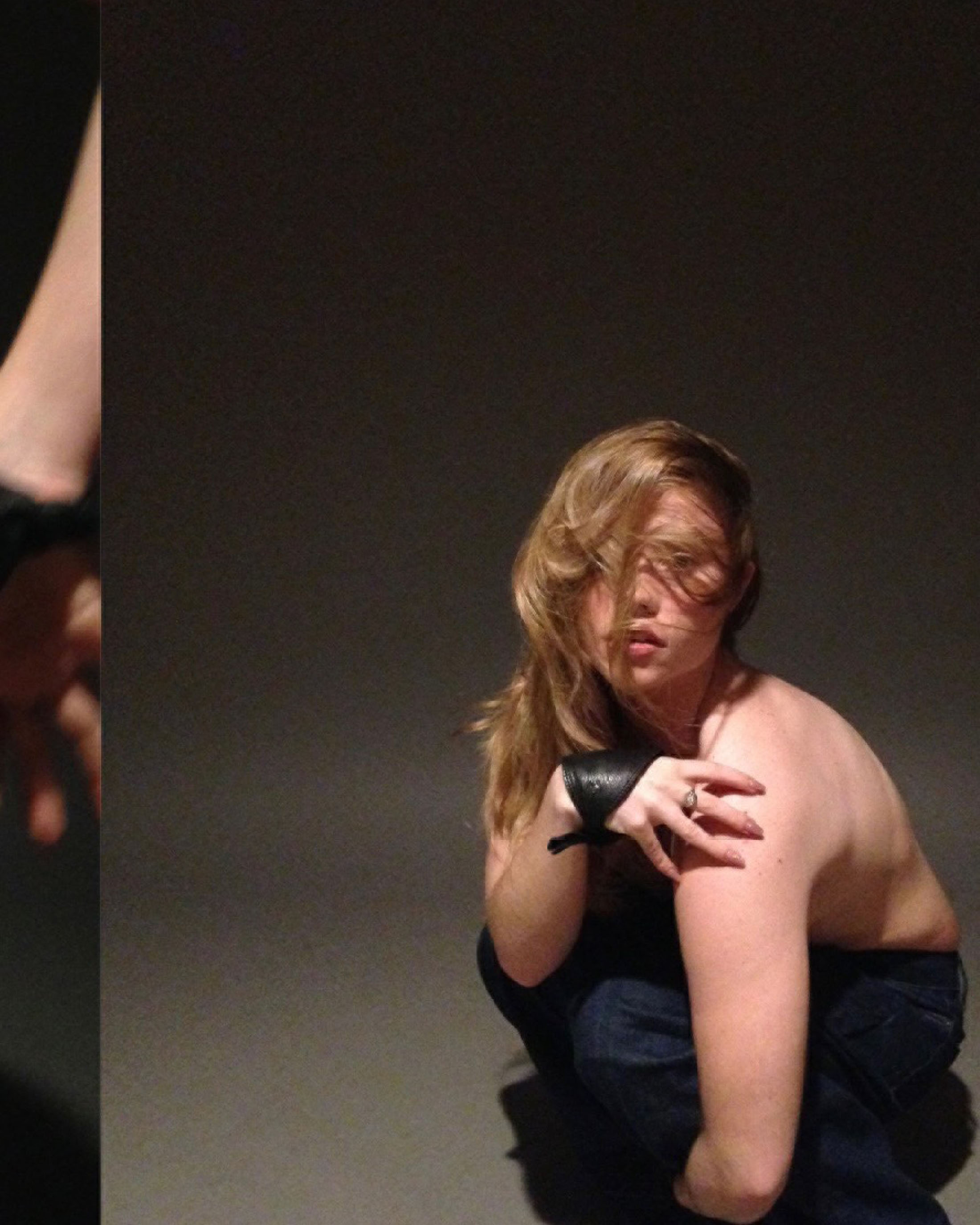 A topless young woman with light brown hair and blue jeans is crouching and looking into the camera in a dark photo studio.