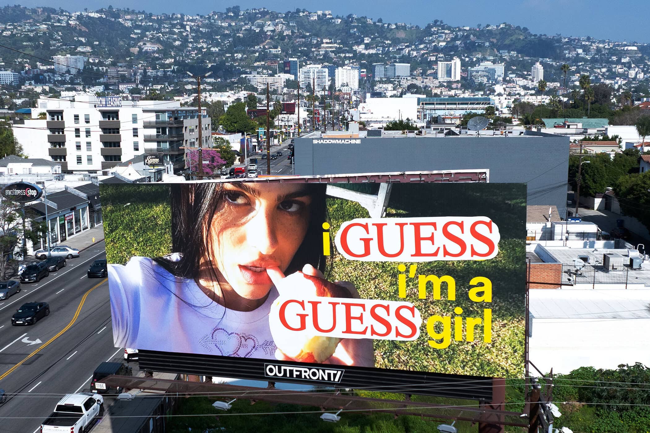 Billboard ad features a young woman in a white shirt with bold text overlay against an urban landscape, promoting a 'GUESS' campaign.